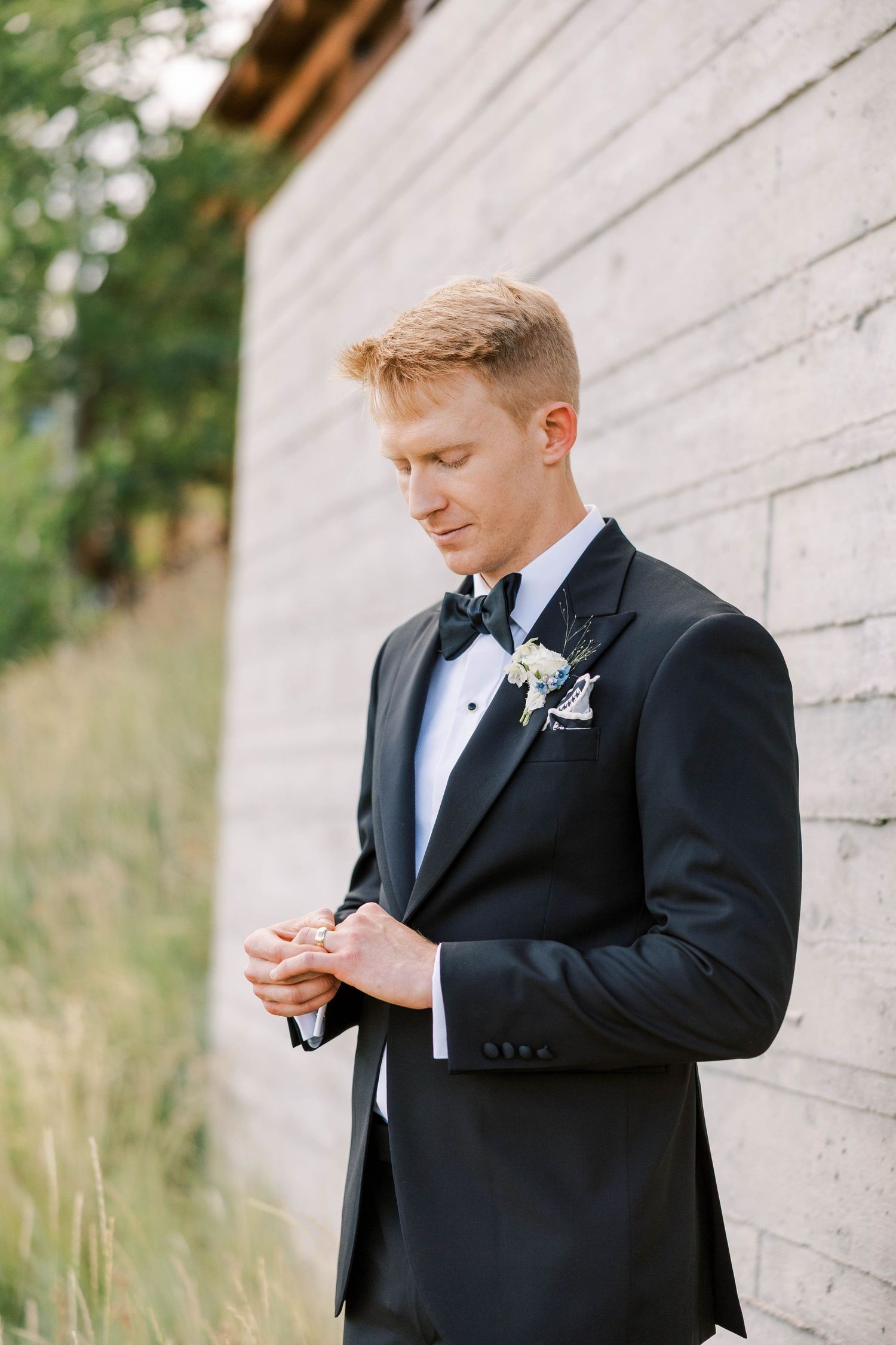Groom in formal attire preparing for ceremony against white wall backdrop.
