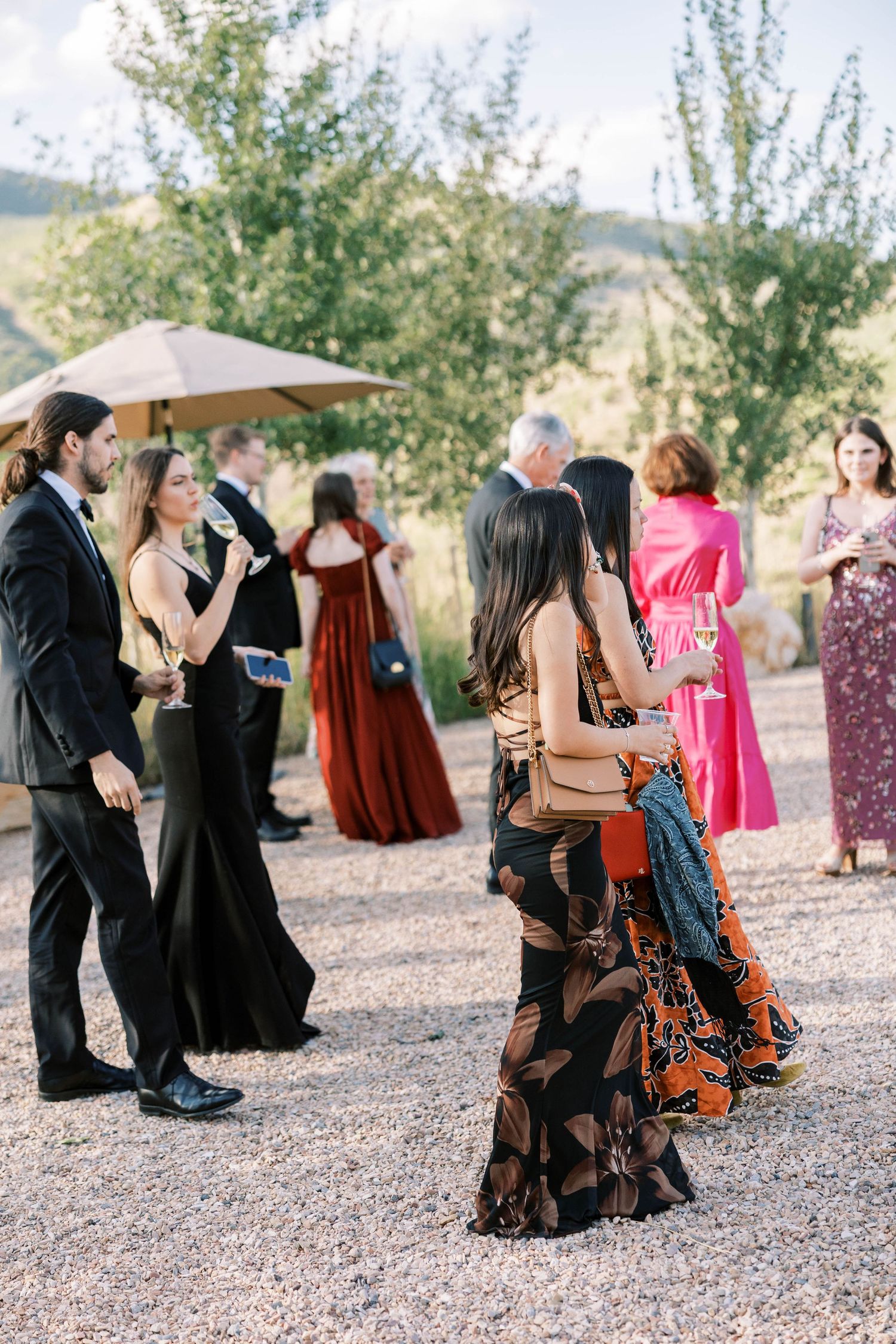 Wedding guests in colorful dresses mingle on a gravel path during outdoor reception.