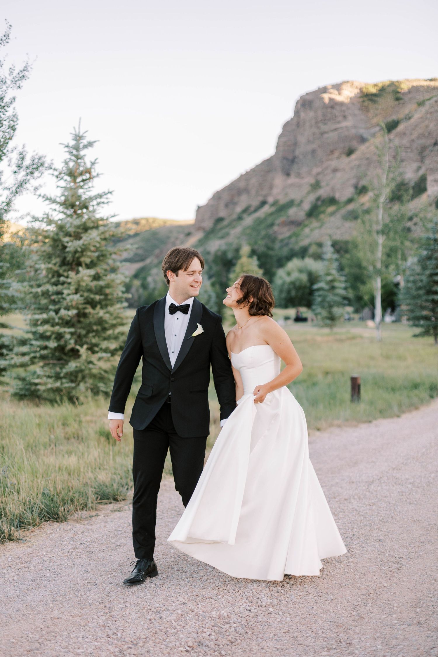 A couple in wedding attire shares a romantic moment on a path surrounded by evergreen trees and mountain scenery.