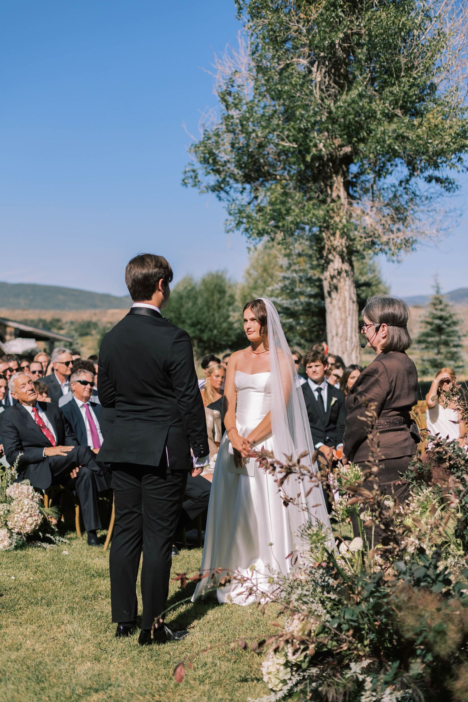A wedding ceremony takes place outdoors on a sunny day with guests gathered on a grassy lawn surrounded by trees.
