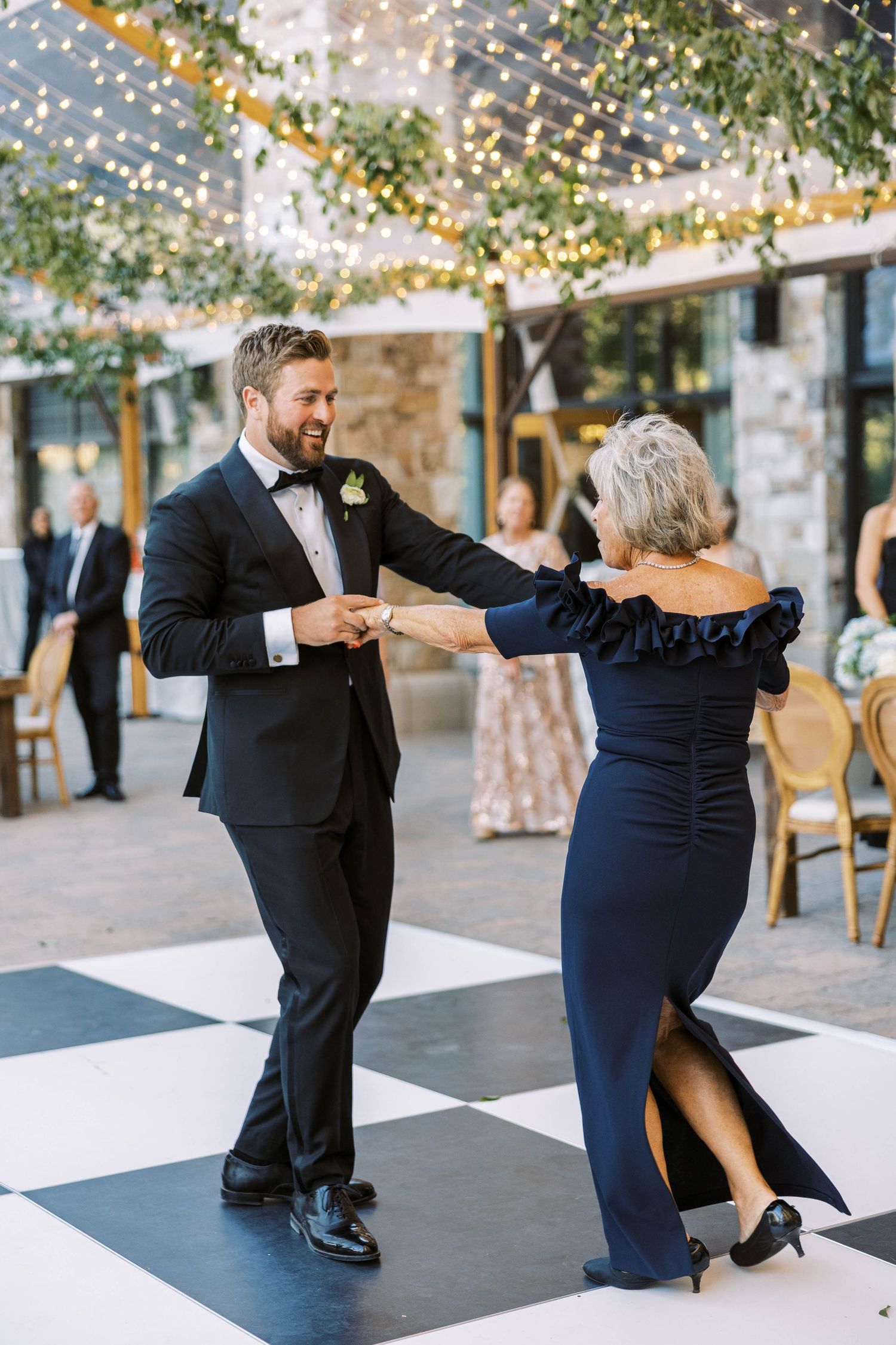 Couple dancing together on a black and white checkered dance floor under twinkling lights at an outdoor event.