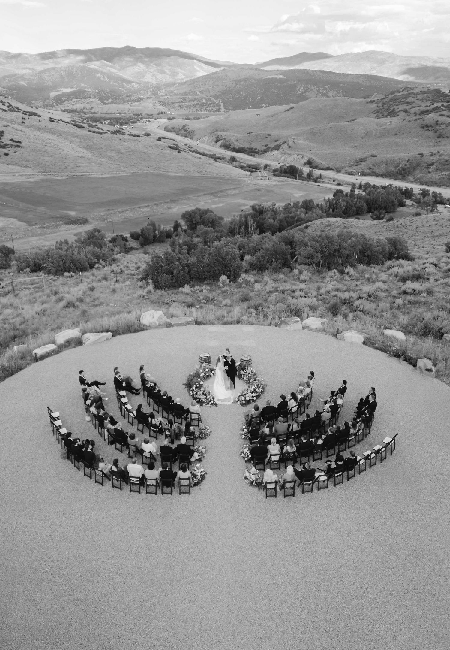 Black and white panorama of outdoor wedding ceremony seating arranged in curved rows with mountain landscape backdrop.