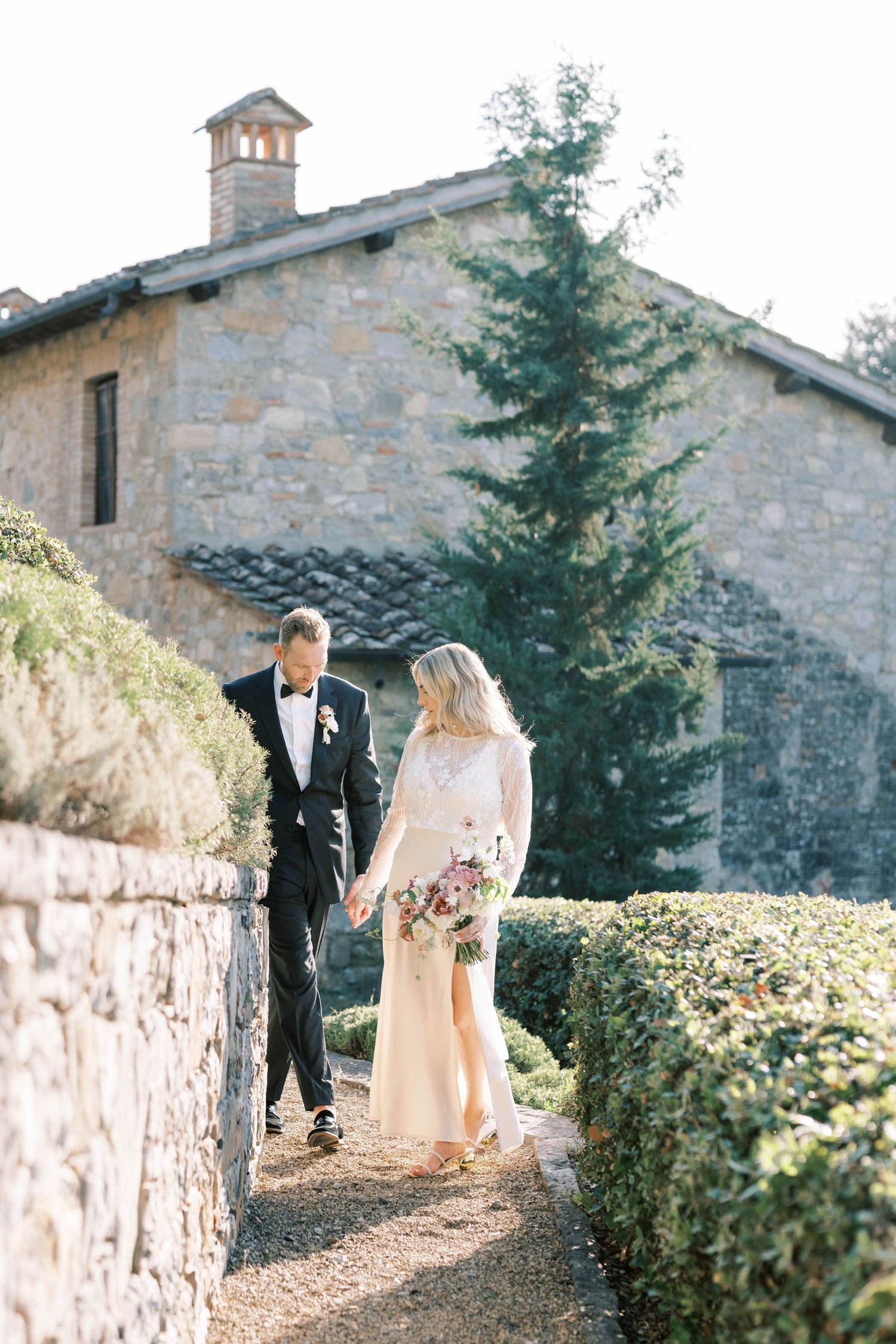 Bride and groom walk hand in hand through a manicured garden path beside a rustic stone villa in Tuscany.