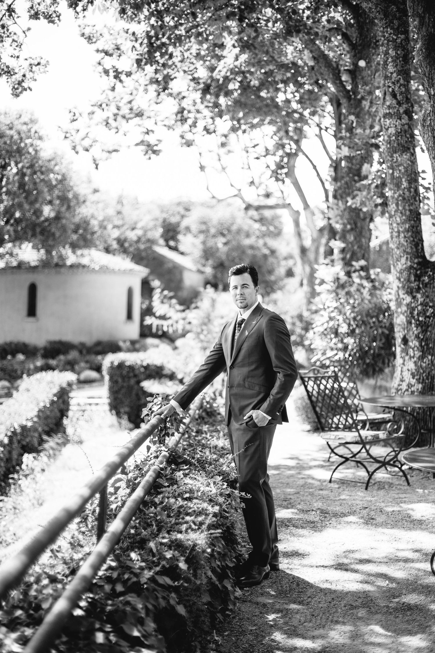 A figure in a dark suit stands elegantly in a garden pathway beside ornate railings.