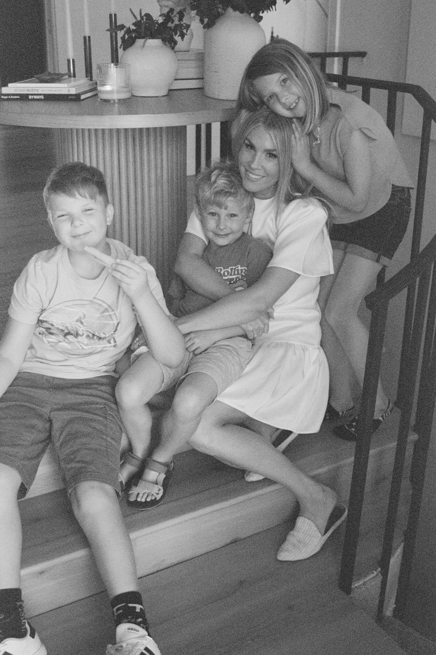 Black and white photo of a smiling woman sitting on stairs with three young children in a cozy home interior.
