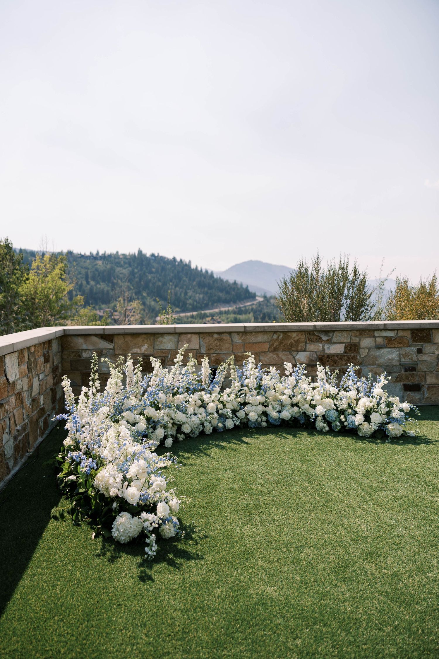 Elegant white floral arrangements line a stone wall overlooking mountain views at an outdoor wedding ceremony setup.