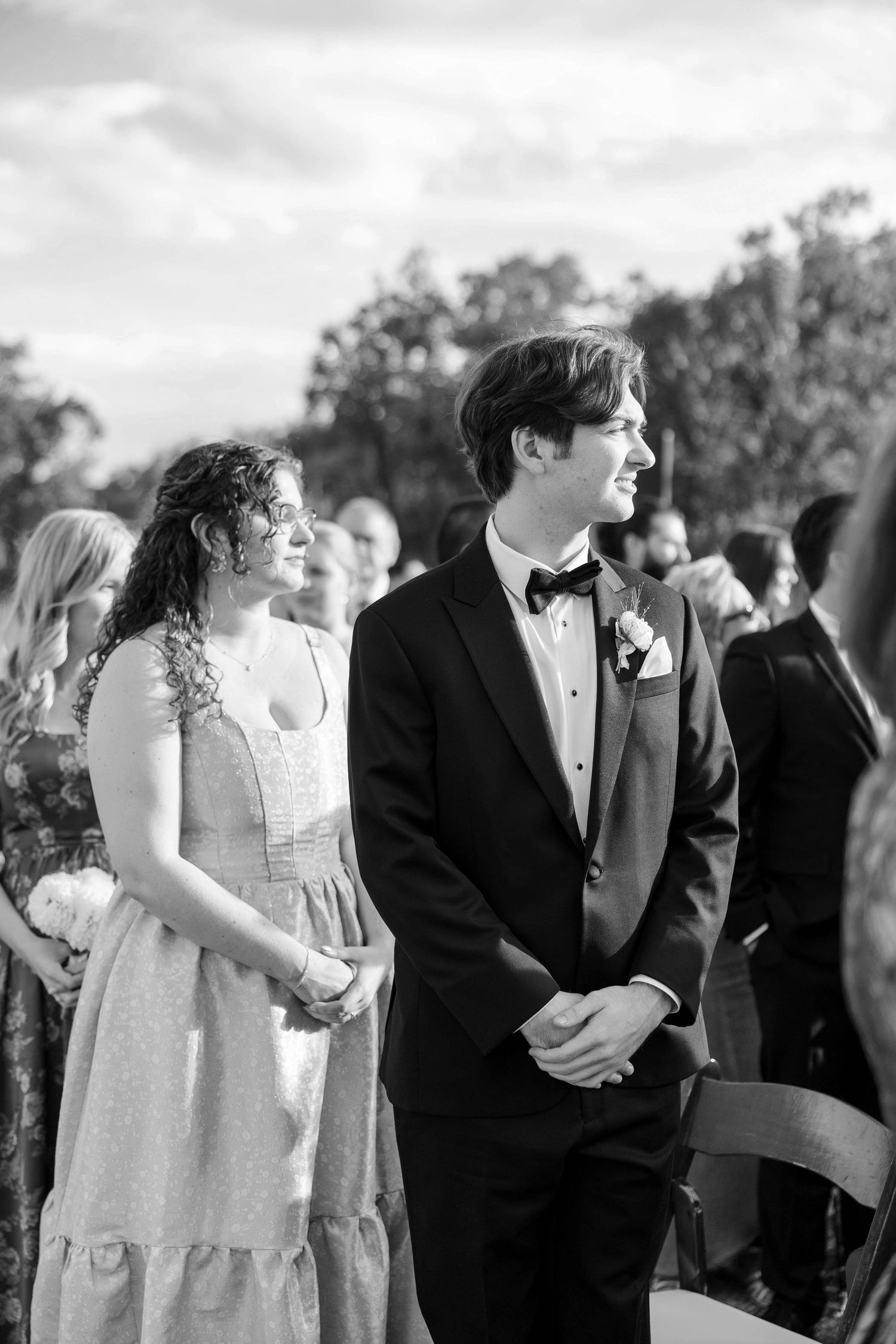 Black and white photograph of wedding guests watching ceremony outdoors under trees.
