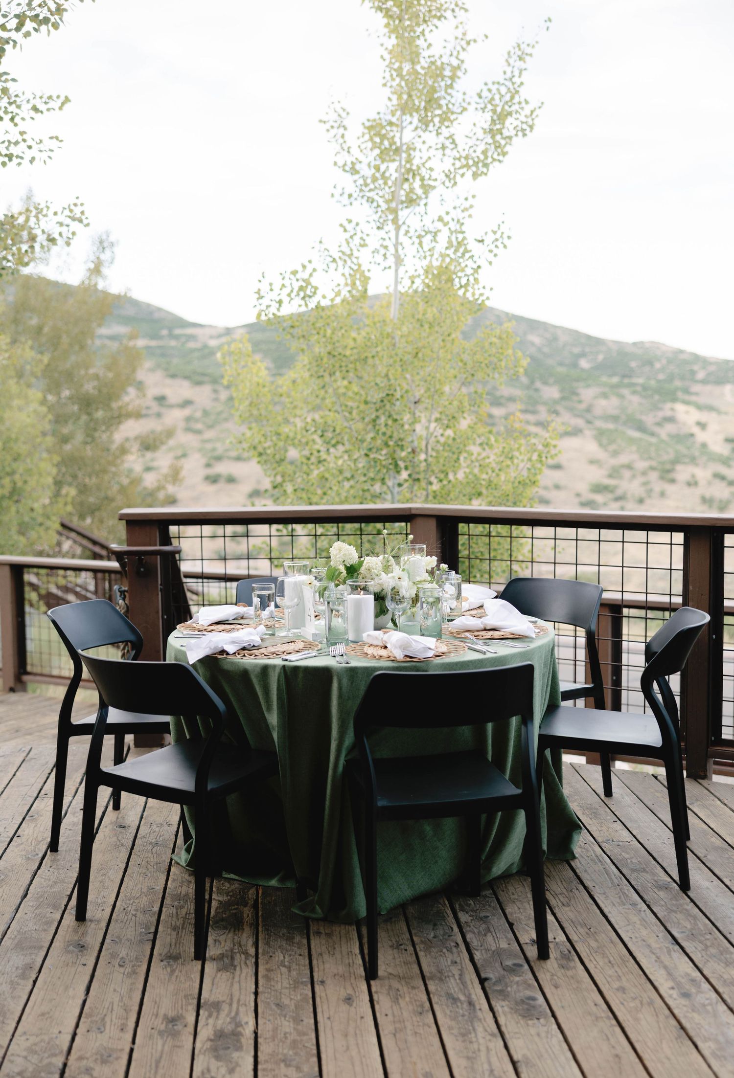 An elegant table setting with dark green tablecloth and modern chairs on a wooden deck overlooking mountains.