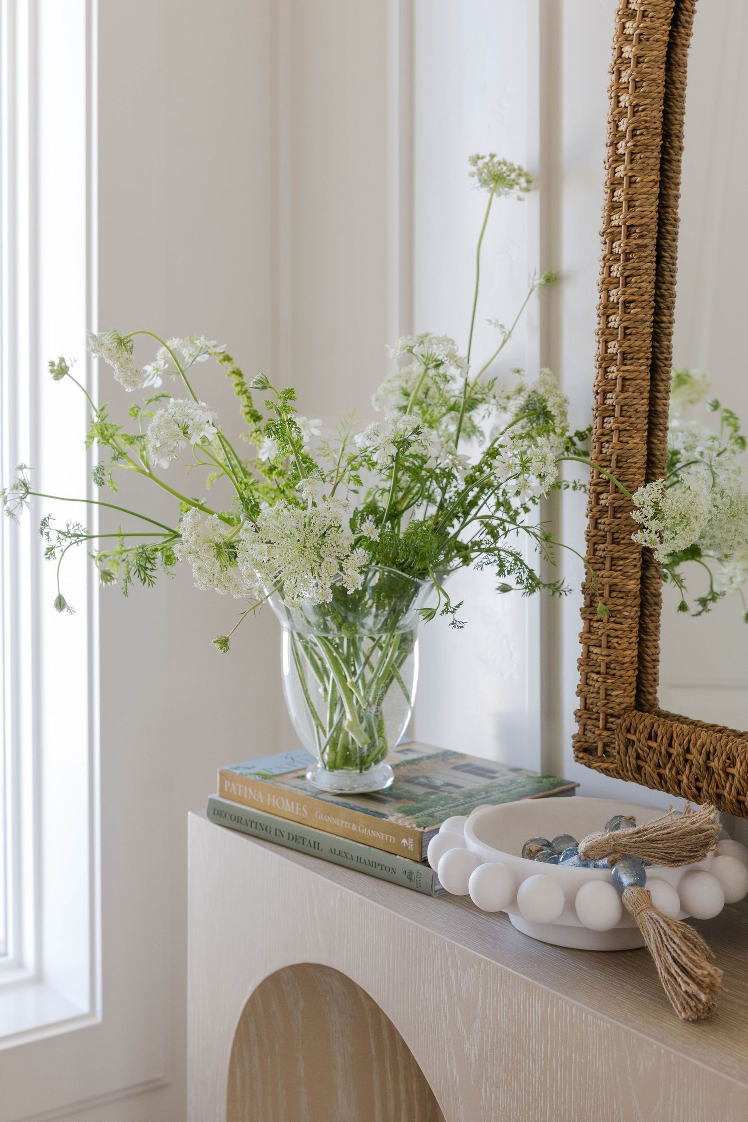 White wildflowers in a glass vase on stacked books beside a decorative bowl on a cream console table with a woven mirror.