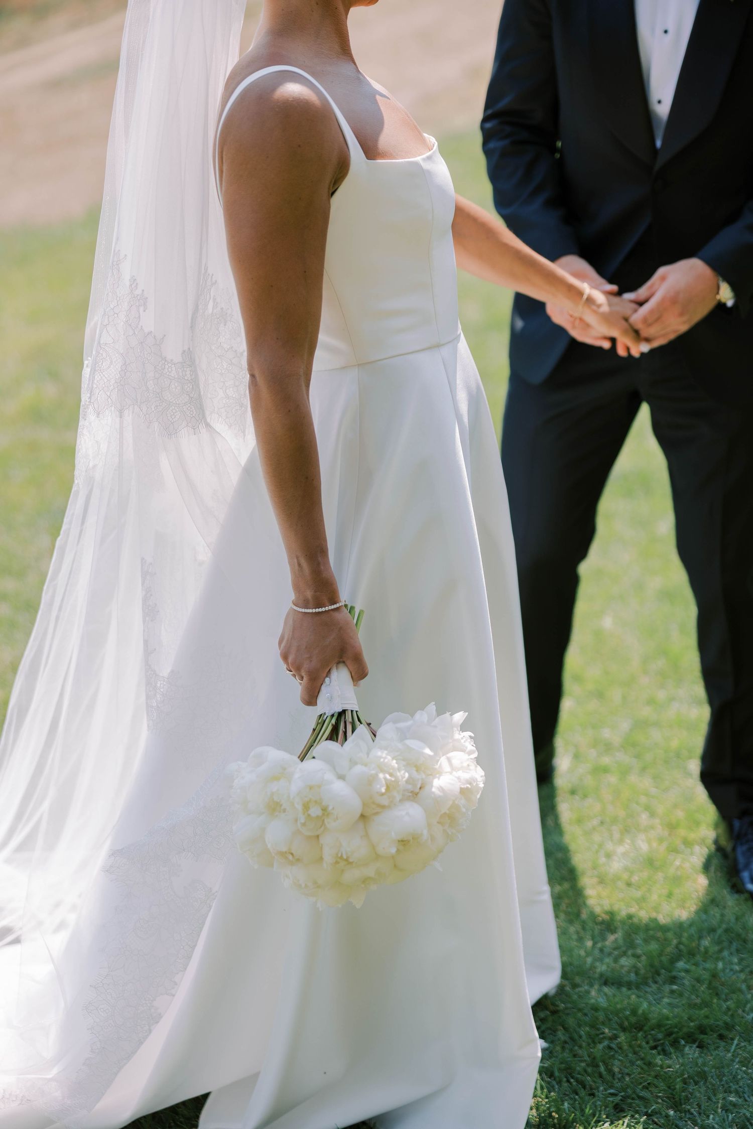 Elegant white wedding dress details with flowing fabric and white bouquet against a grassy outdoor backdrop.