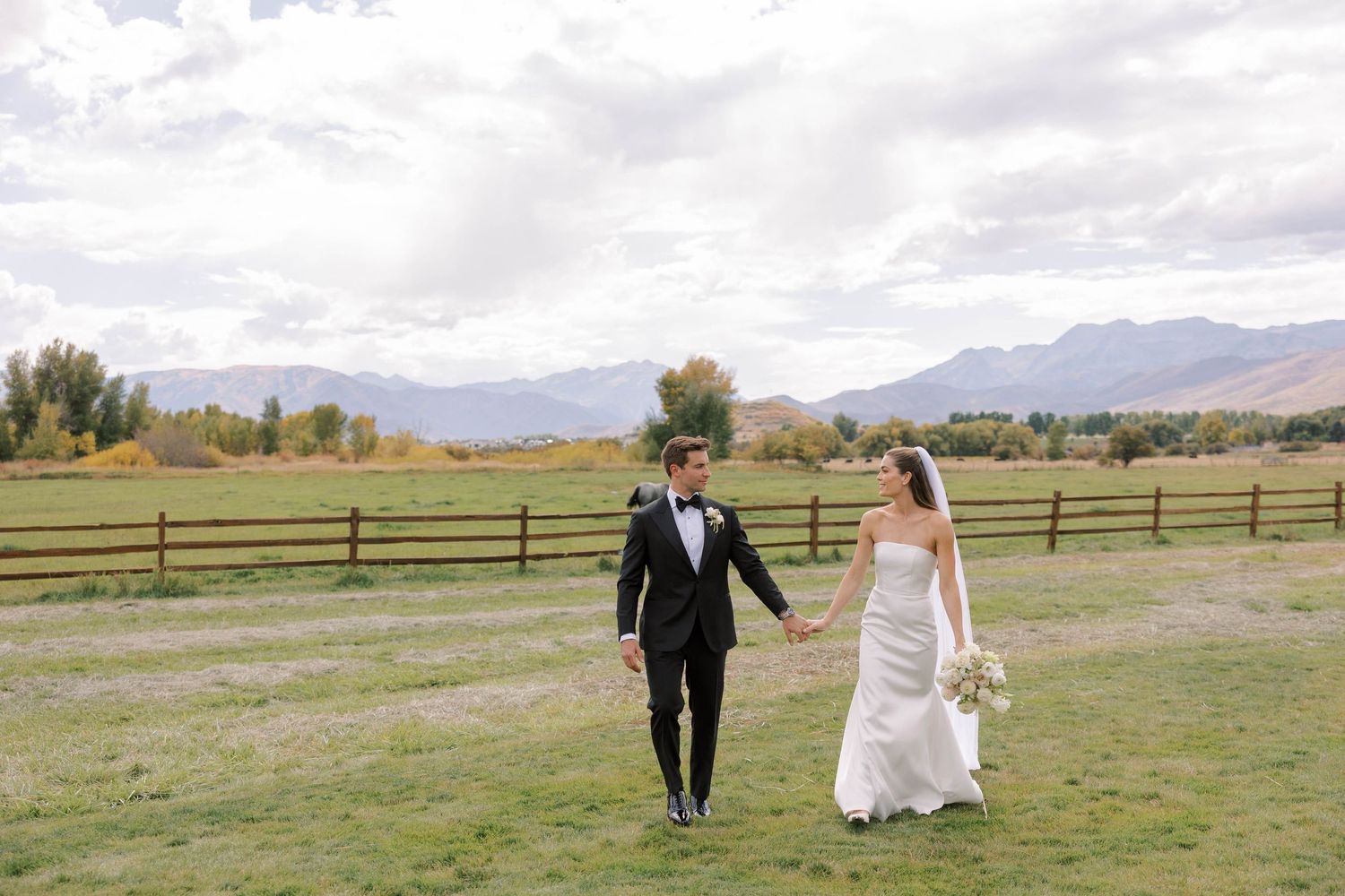 A couple walks through a scenic mountain landscape with rustic wooden fences and autumn foliage in the background.