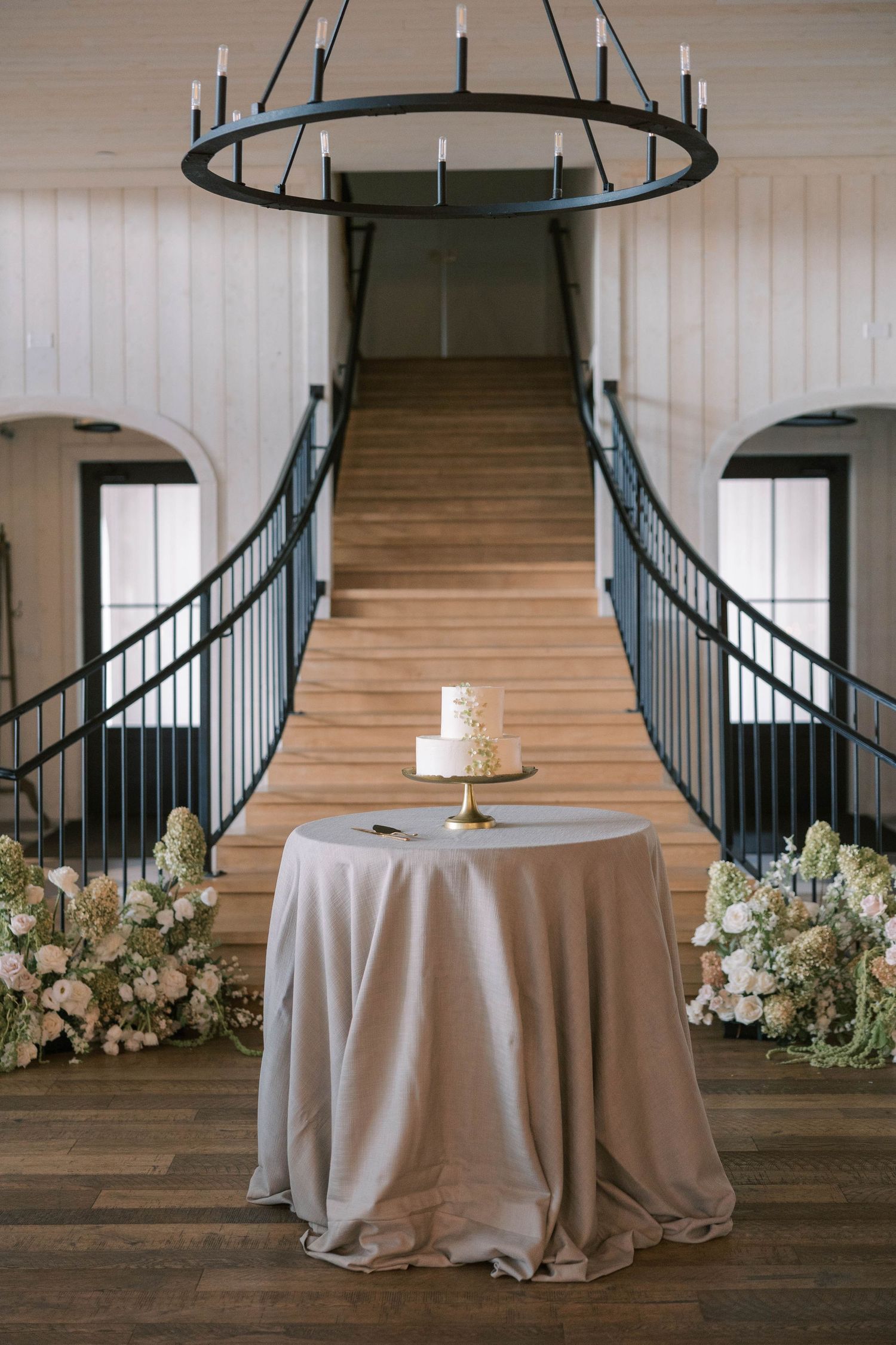 Elegant wedding reception setup with grand staircase, round tables with gray linens, and circular black chandelier.