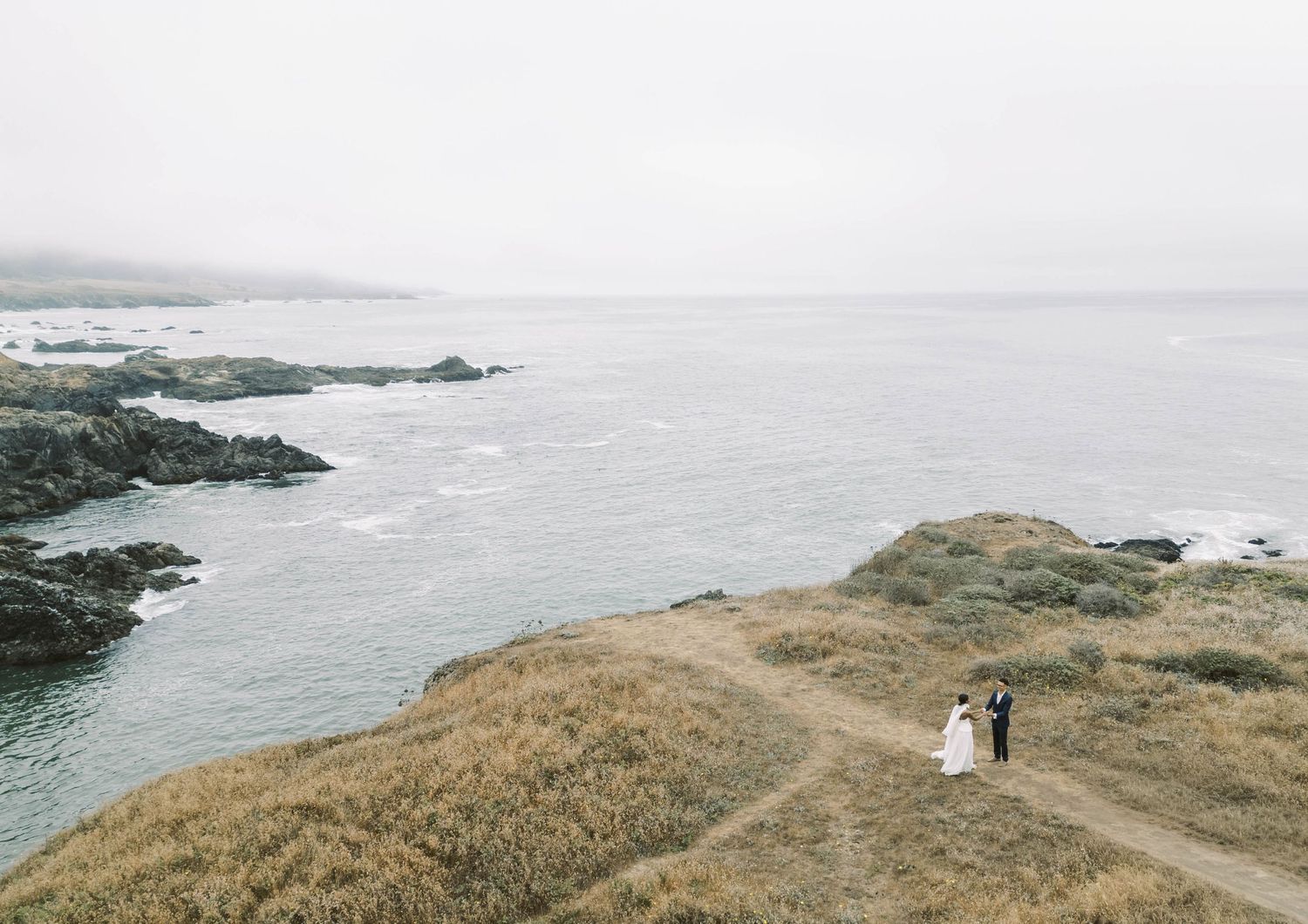 Coastal wedding photography sequence showing a couple walking along rugged cliffs overlooking a misty ocean shoreline.