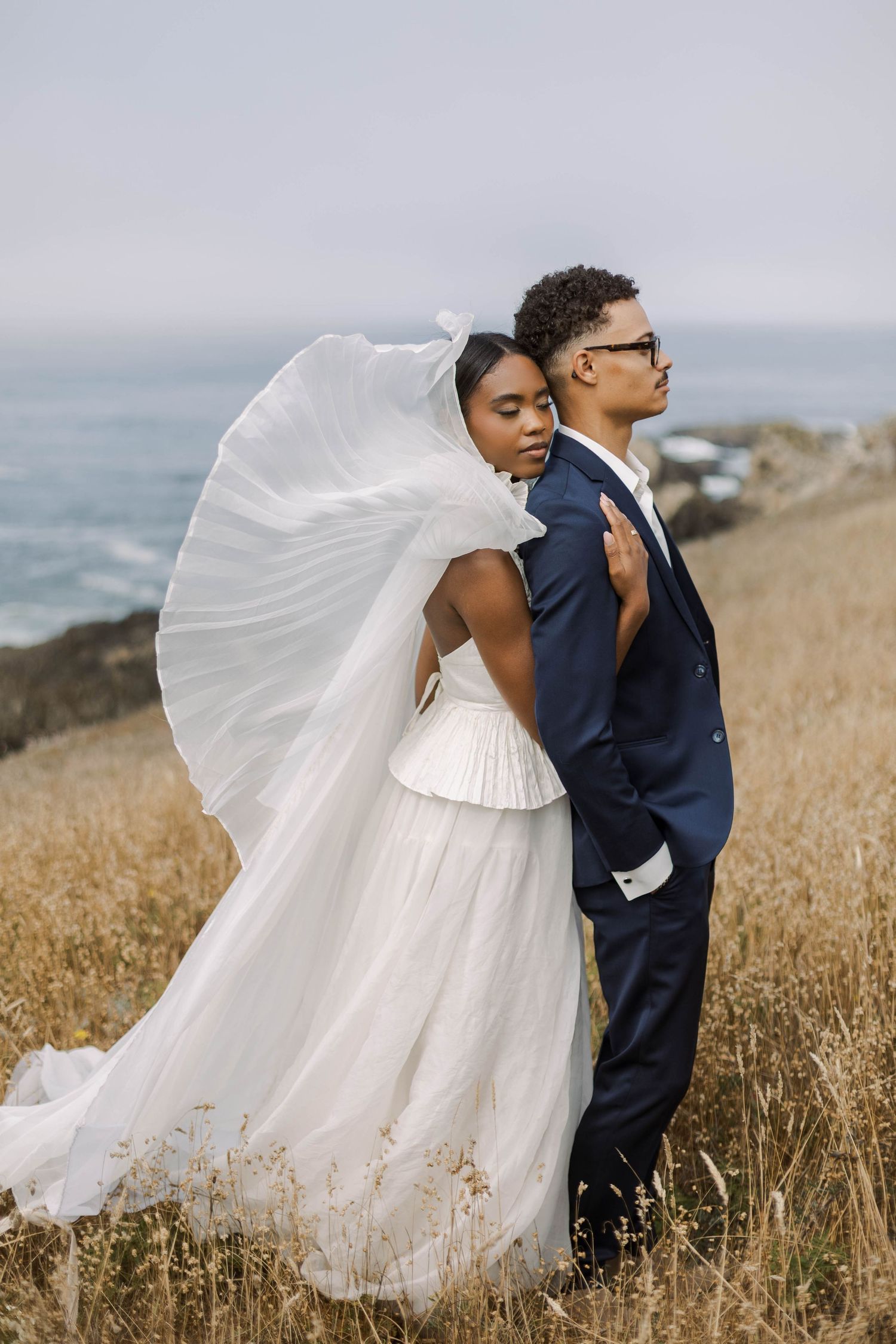 A couple in wedding attire stands on a coastal cliff with flowing veil and tall grass at sunset.