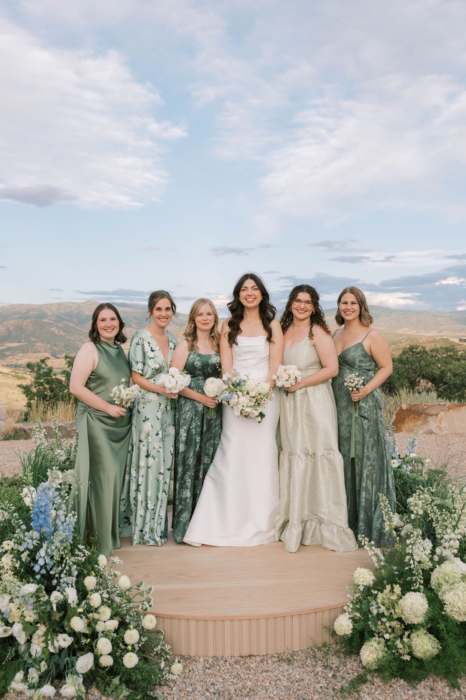 A wedding party in sage green and neutral dresses poses outdoors against a mountain backdrop.