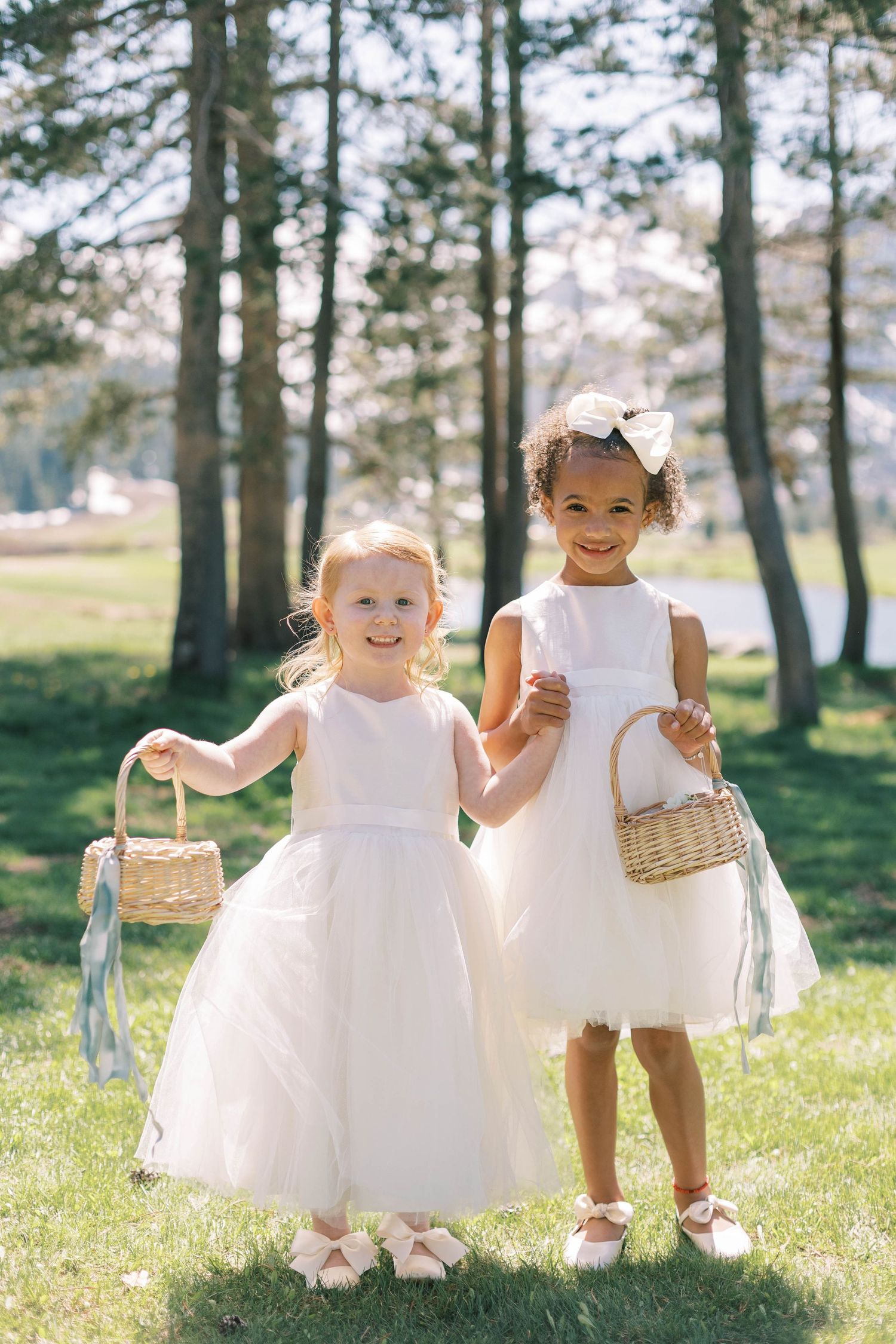 Two flower girls in white dresses hold baskets while standing on grass with trees in background.