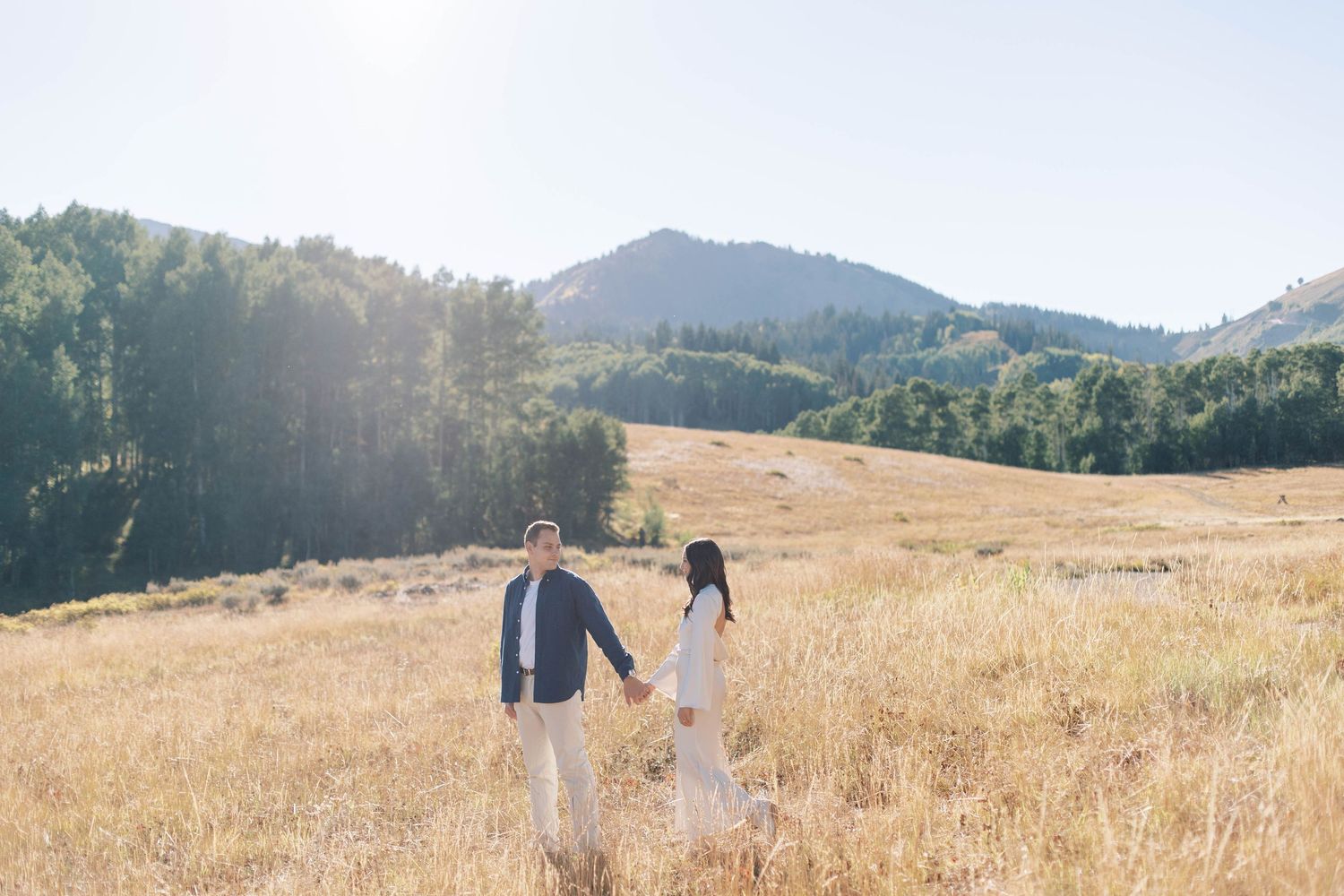 Couple holding hands walking through golden mountain meadow at sunset.