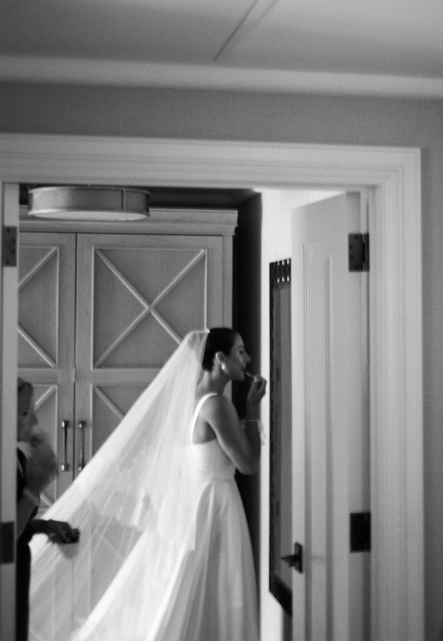 A bride in a flowing white wedding gown and veil stands in a doorway in this black and white photograph.