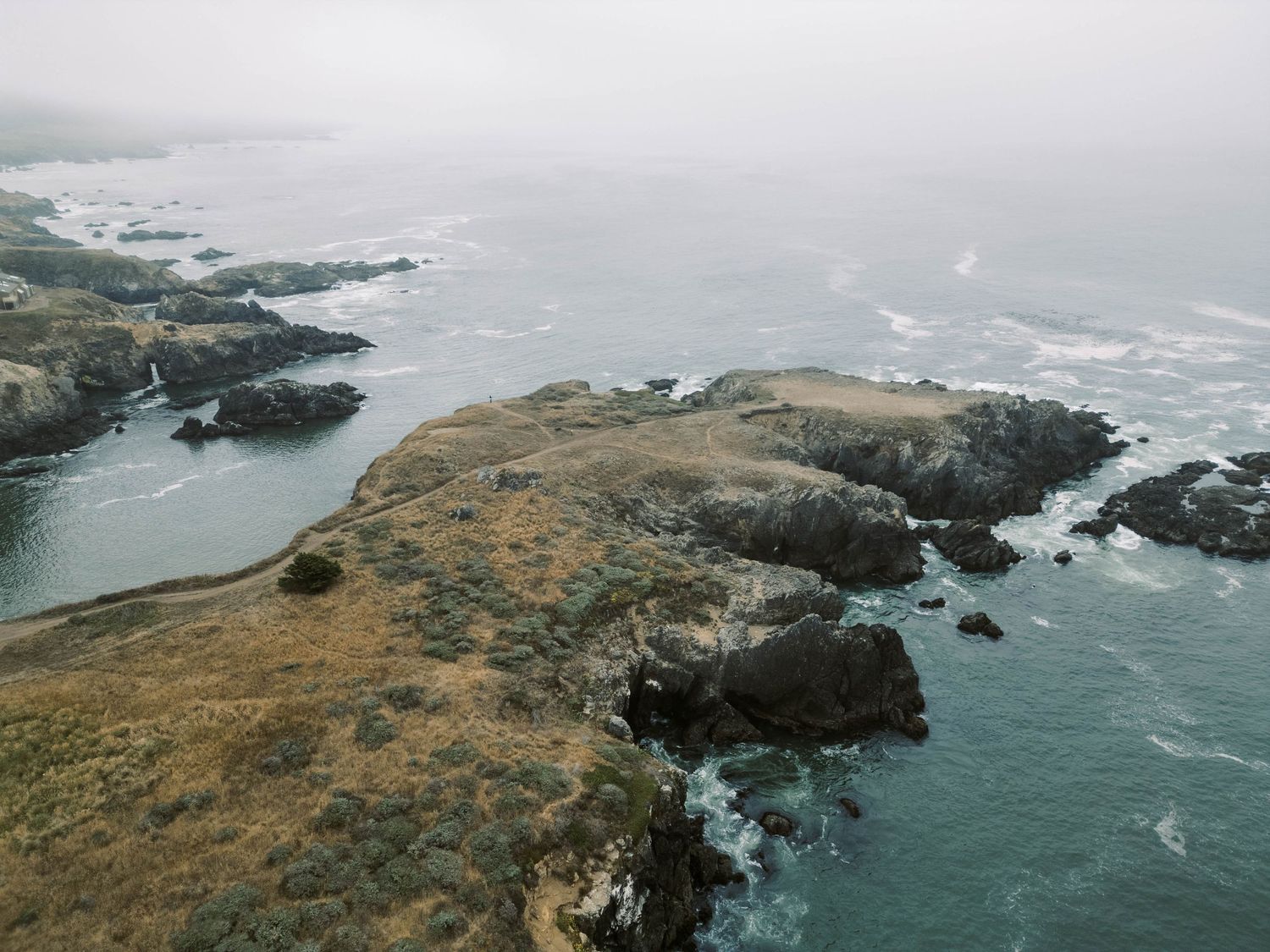 Dramatic aerial view of rugged coastal cliffs and crashing waves along a rocky shoreline on a misty day.