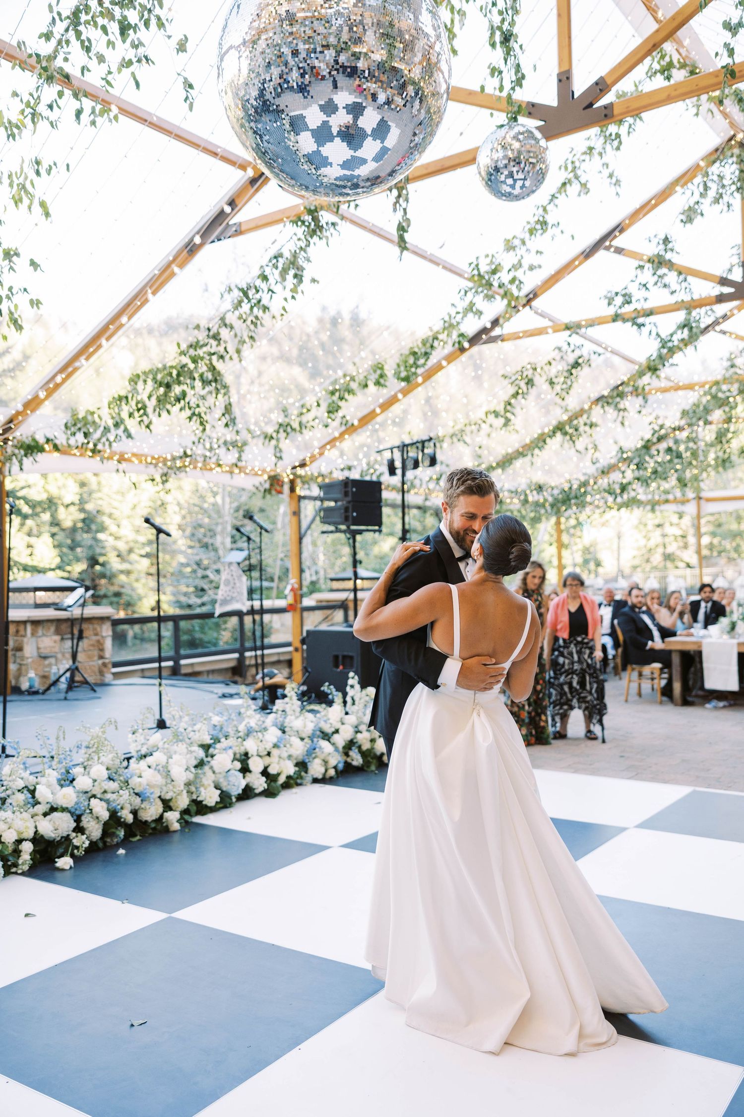 A wedding reception setup in a clear tent with hanging greenery, disco balls, and a white dance floor for celebrations.
