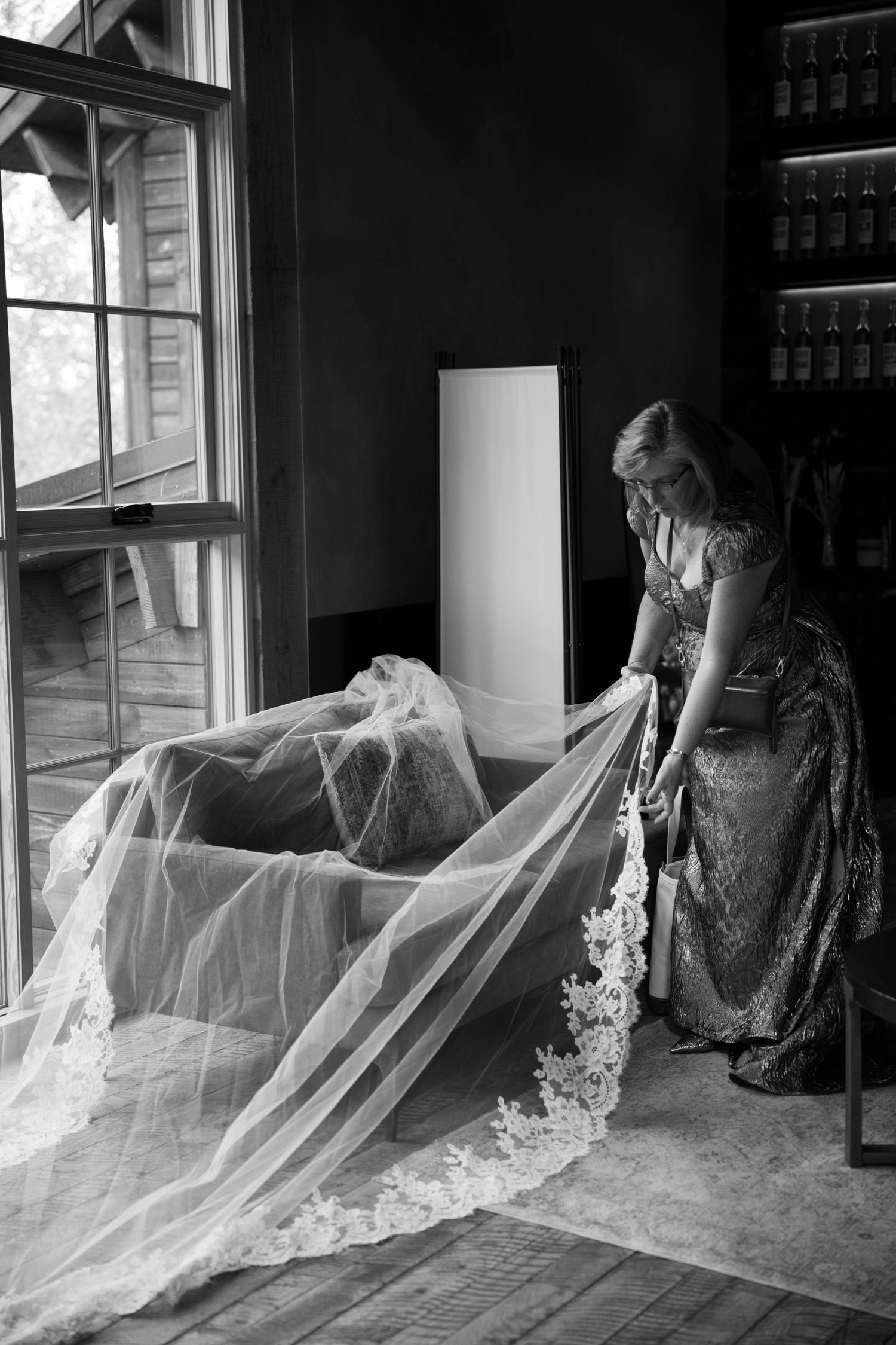 Flowing white wedding veil being adjusted near windows in dark room.