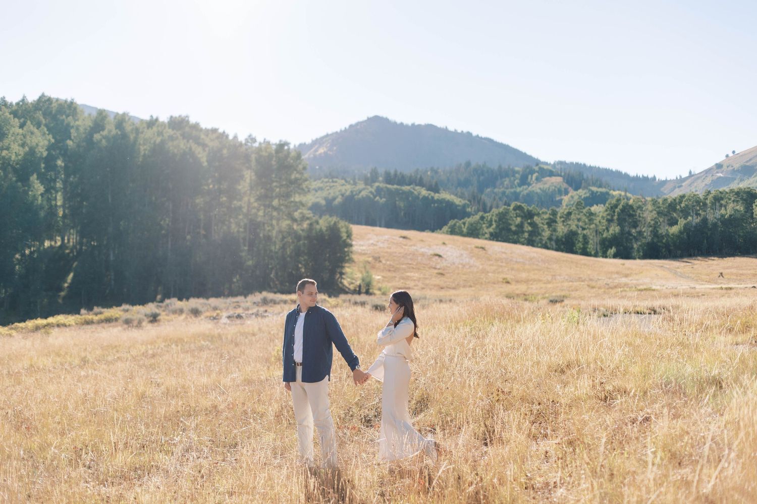 Couple strolling through sun-drenched mountain meadow holding hands.
