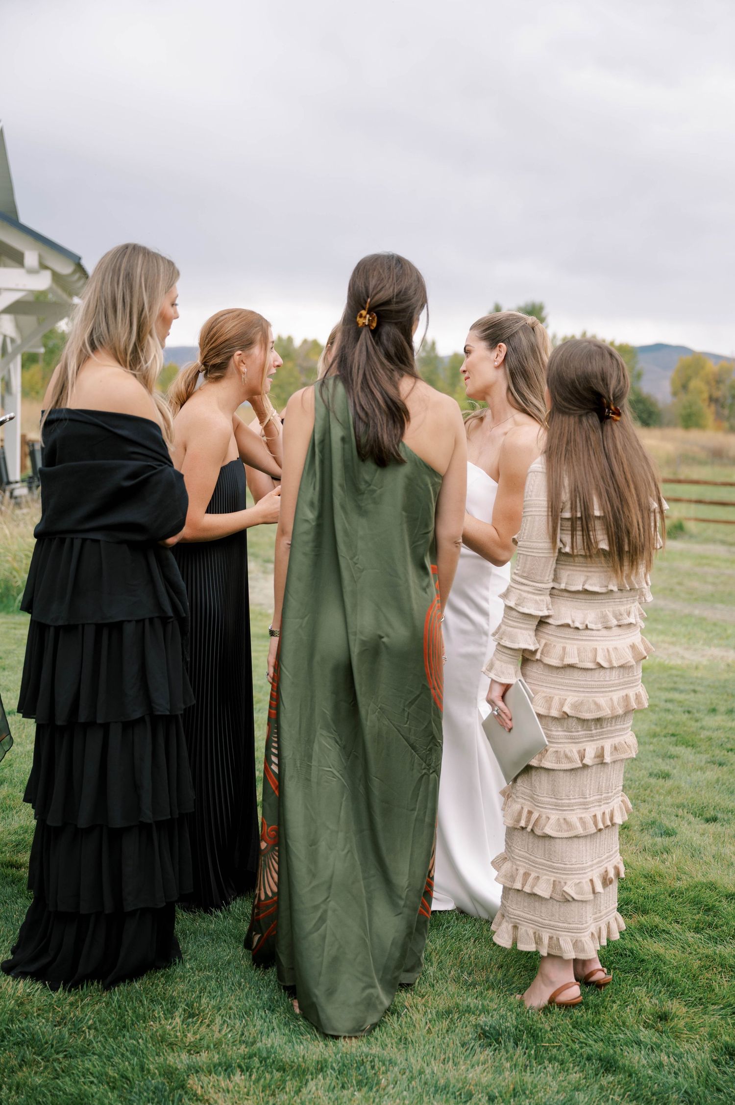 Four women in long dresses stand together in a field taking photos on a cloudy day.