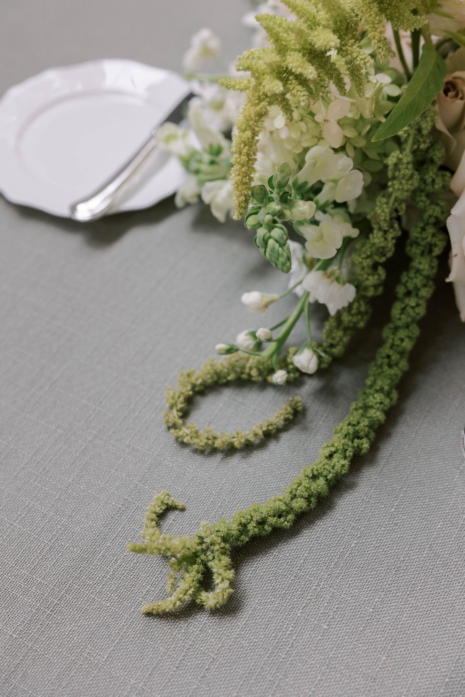 Green amaranthus flowers and white blooms cascade elegantly across a gray linen tablecloth beside a white plate.