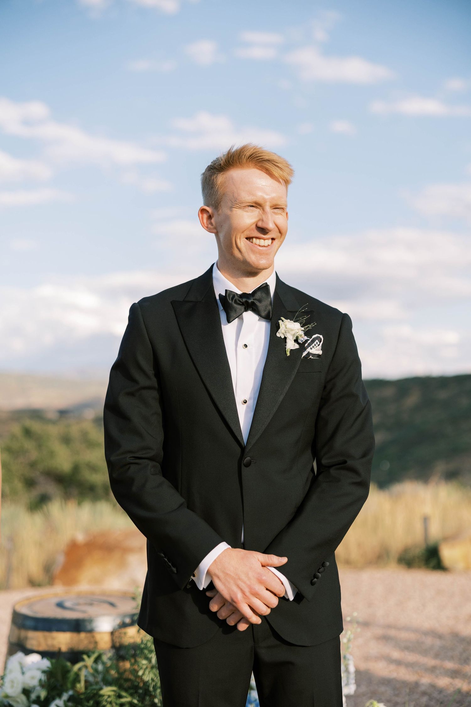 Groom in classic black tuxedo smiles joyfully against blue sky and mountain landscape.