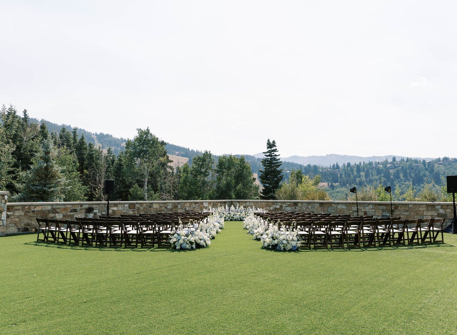 Rows of dark wooden chairs arranged in a curved pattern on a lush green lawn with mountains and trees in the background.