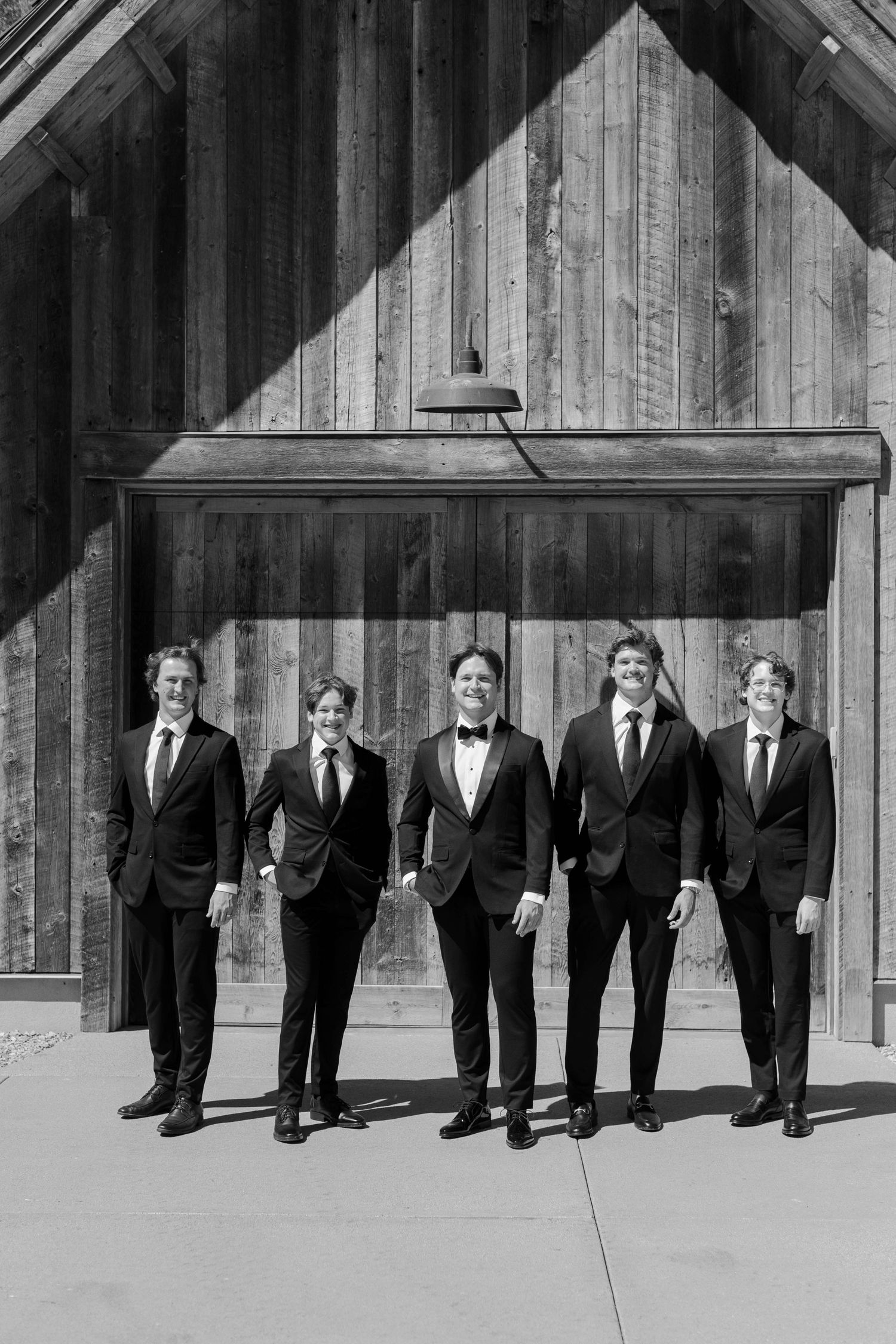 A creative black and white panoramic photo of groomsmen standing in front of a rustic wooden barn wall.