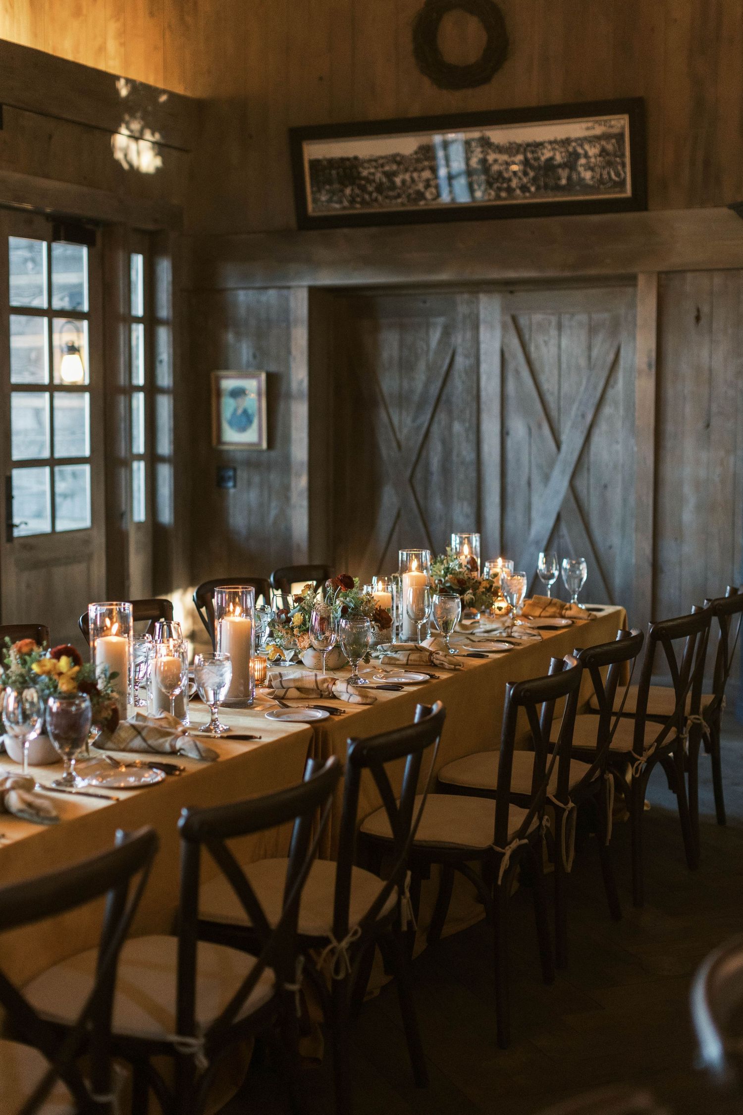 Rustic barn wedding reception with long wooden table and cross-back chairs.