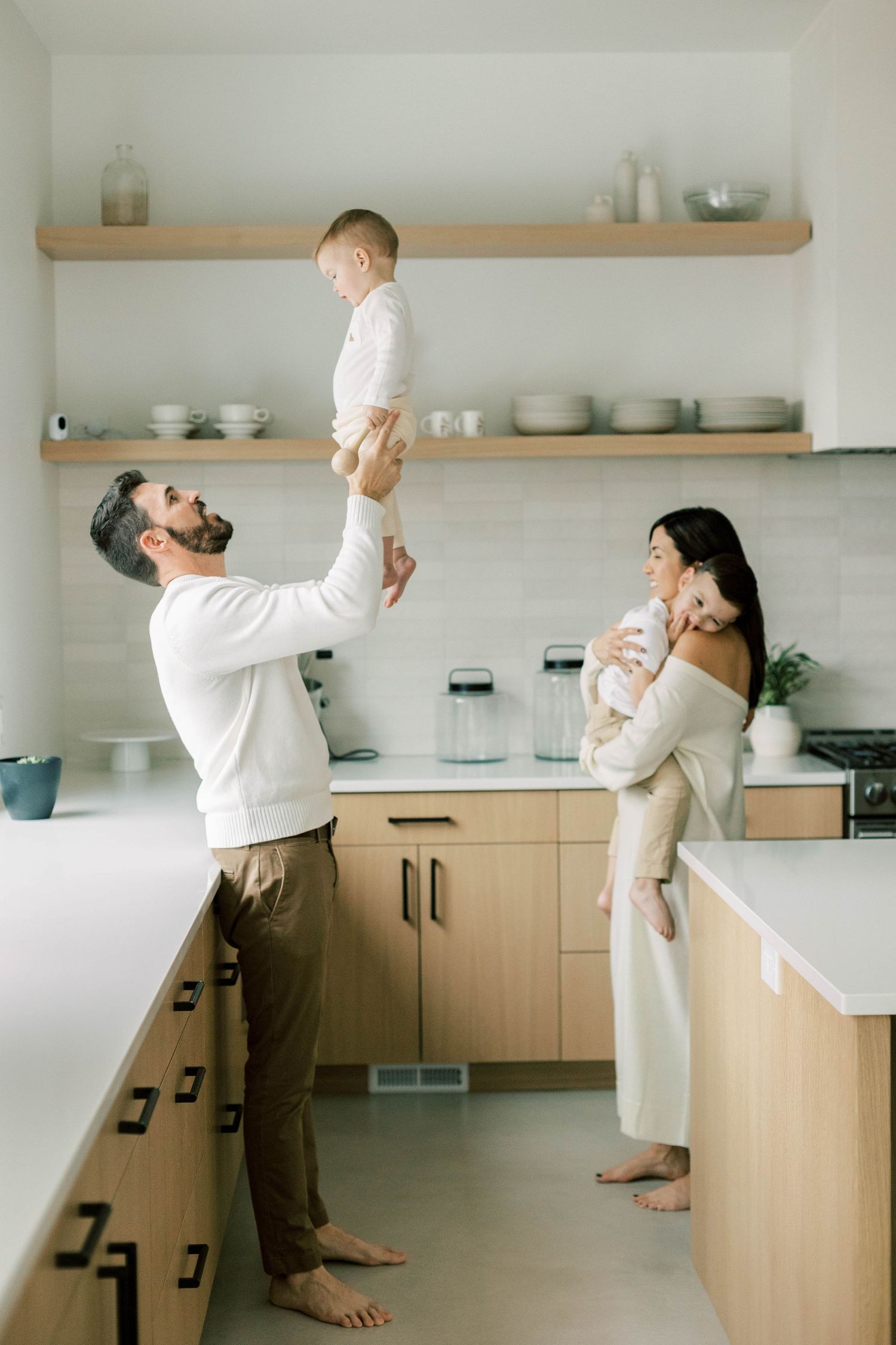 Playful family scene in modern kitchen with light wood cabinets and open shelving.