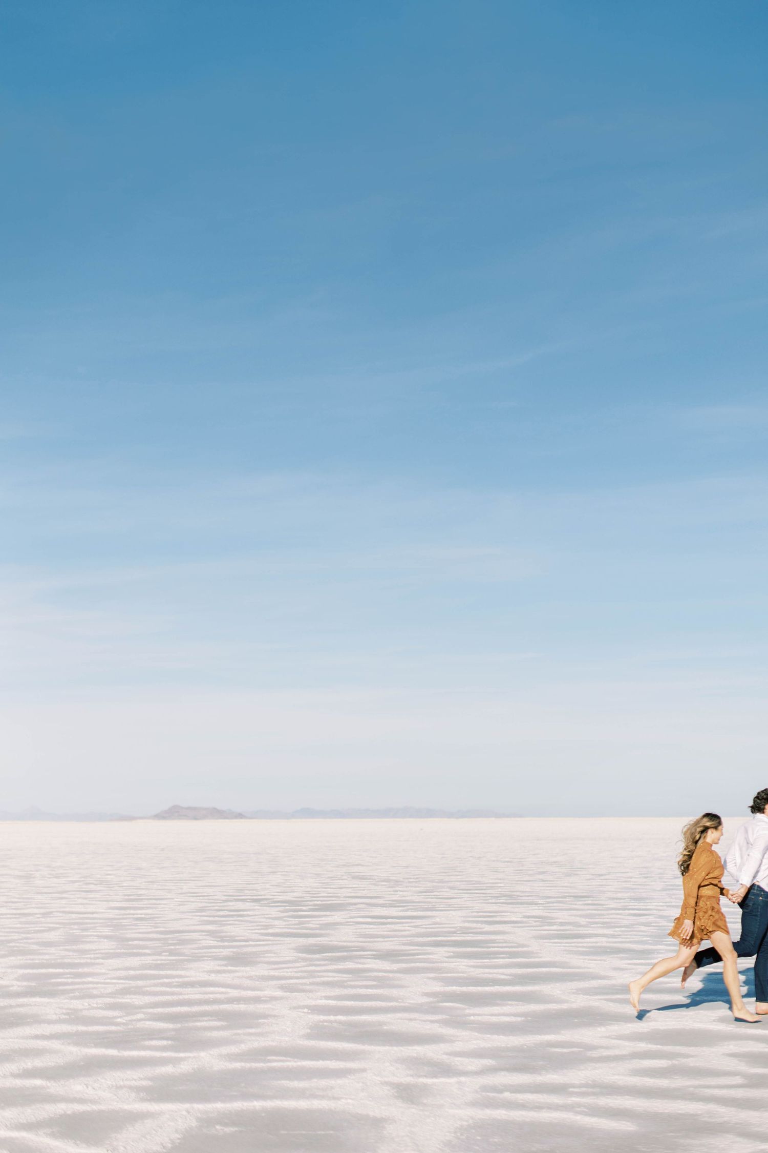 Young couple running playfully across a vast white salt flat under clear blue skies.