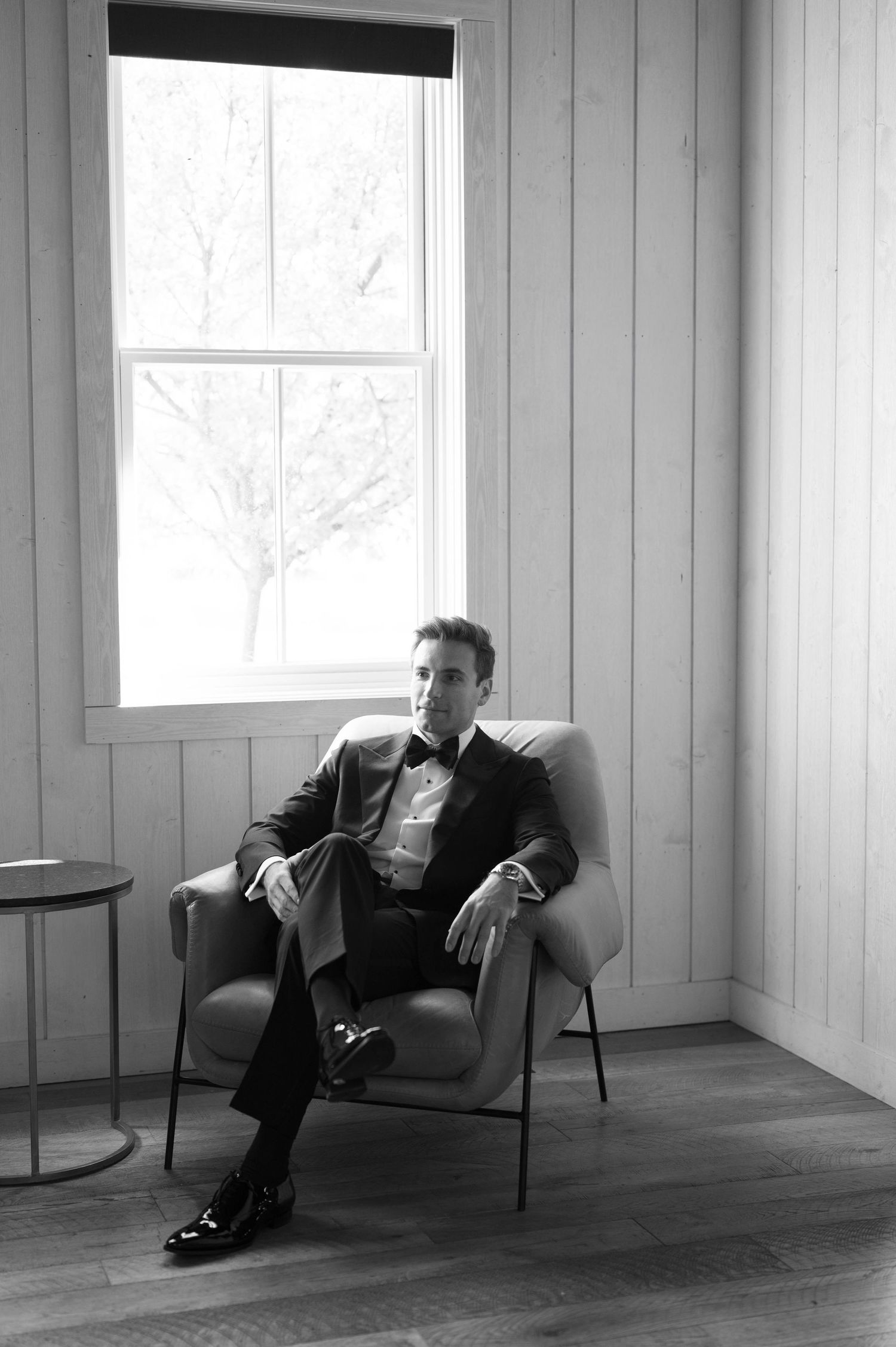 Black and white portrait of person lounging in chair against wooden paneled wall in minimalist room.