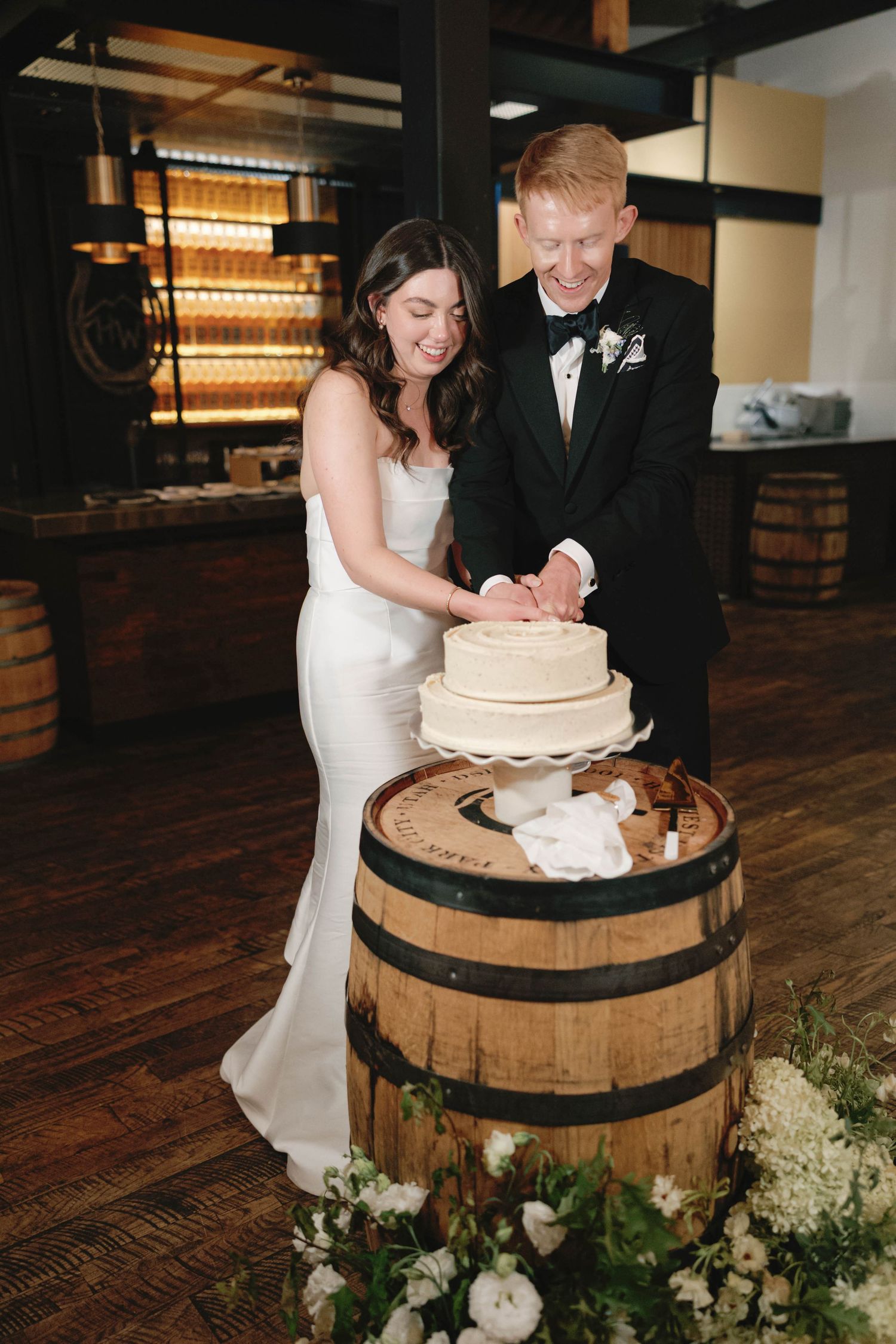 Couple cutting their wedding cake displayed on a rustic wooden barrel decorated with white flowers.