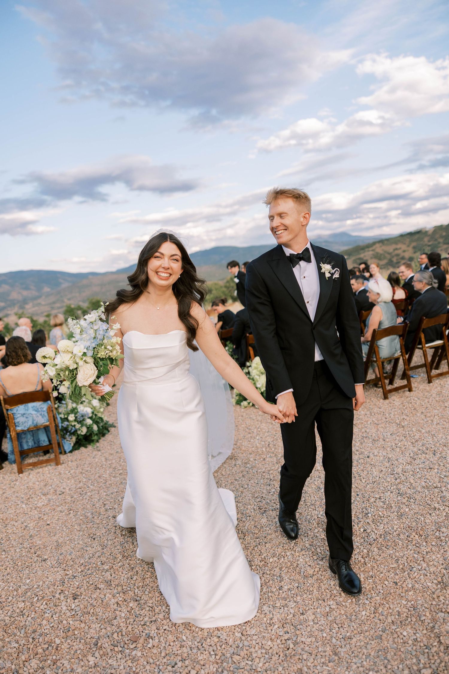 A wedding couple walks hand in hand on a gravel path with mountain views and wooden chairs in the background.