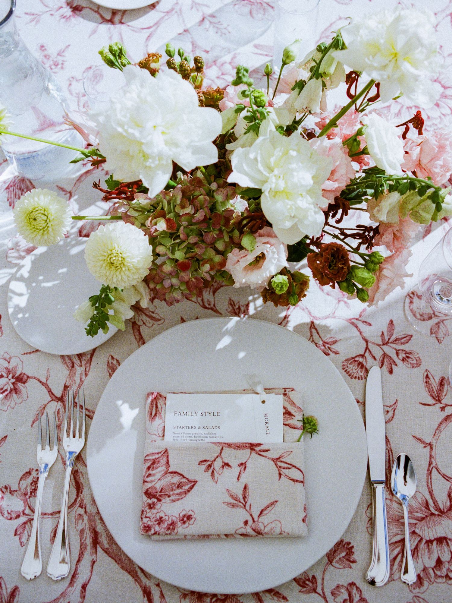 Elegant place setting with white and pink floral decor on pink toile tablecloth featuring delicate china and silverware.