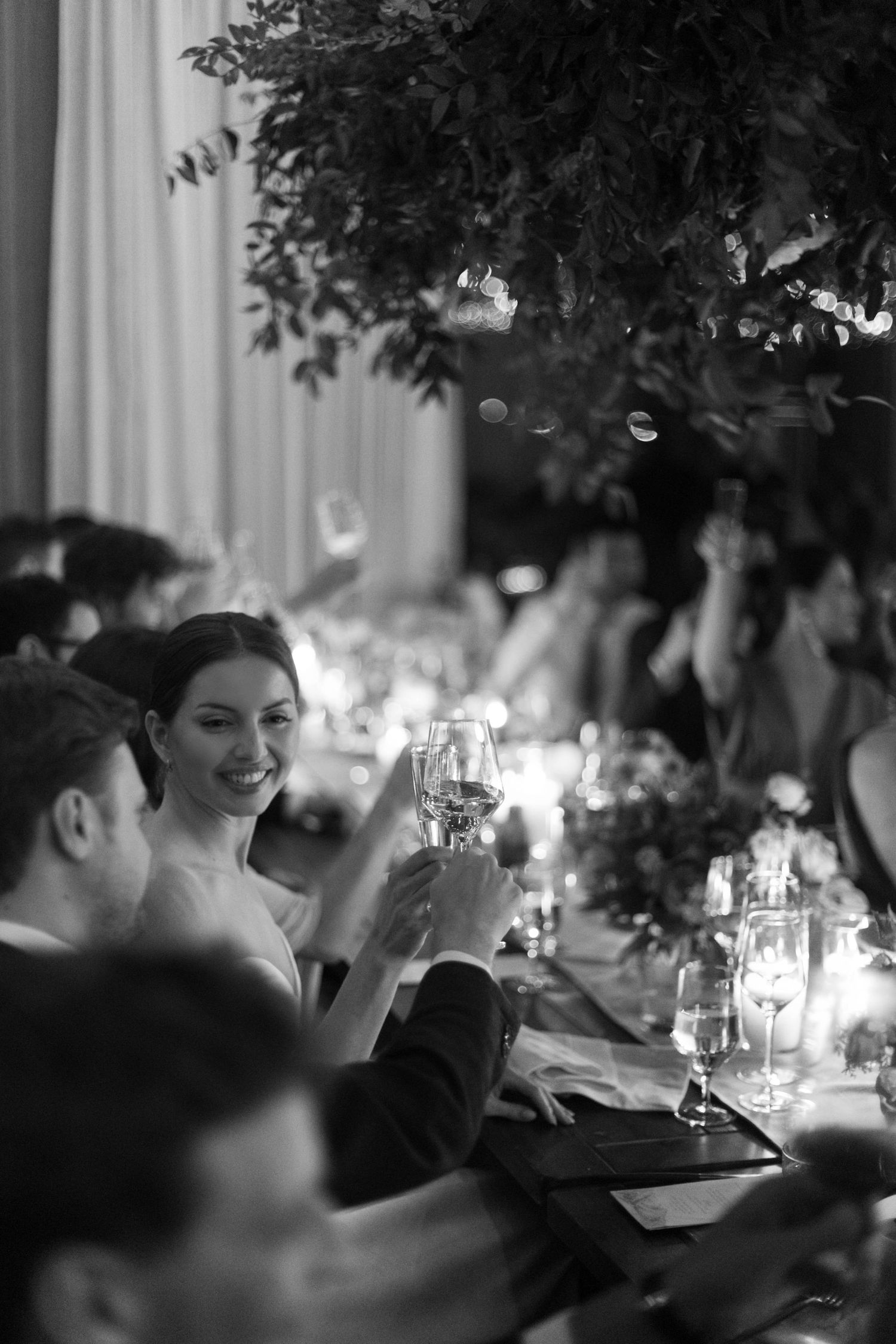 A black and white photo at an elegant dinner party showing guests toasting with wine glasses amid candlelight and foliage.