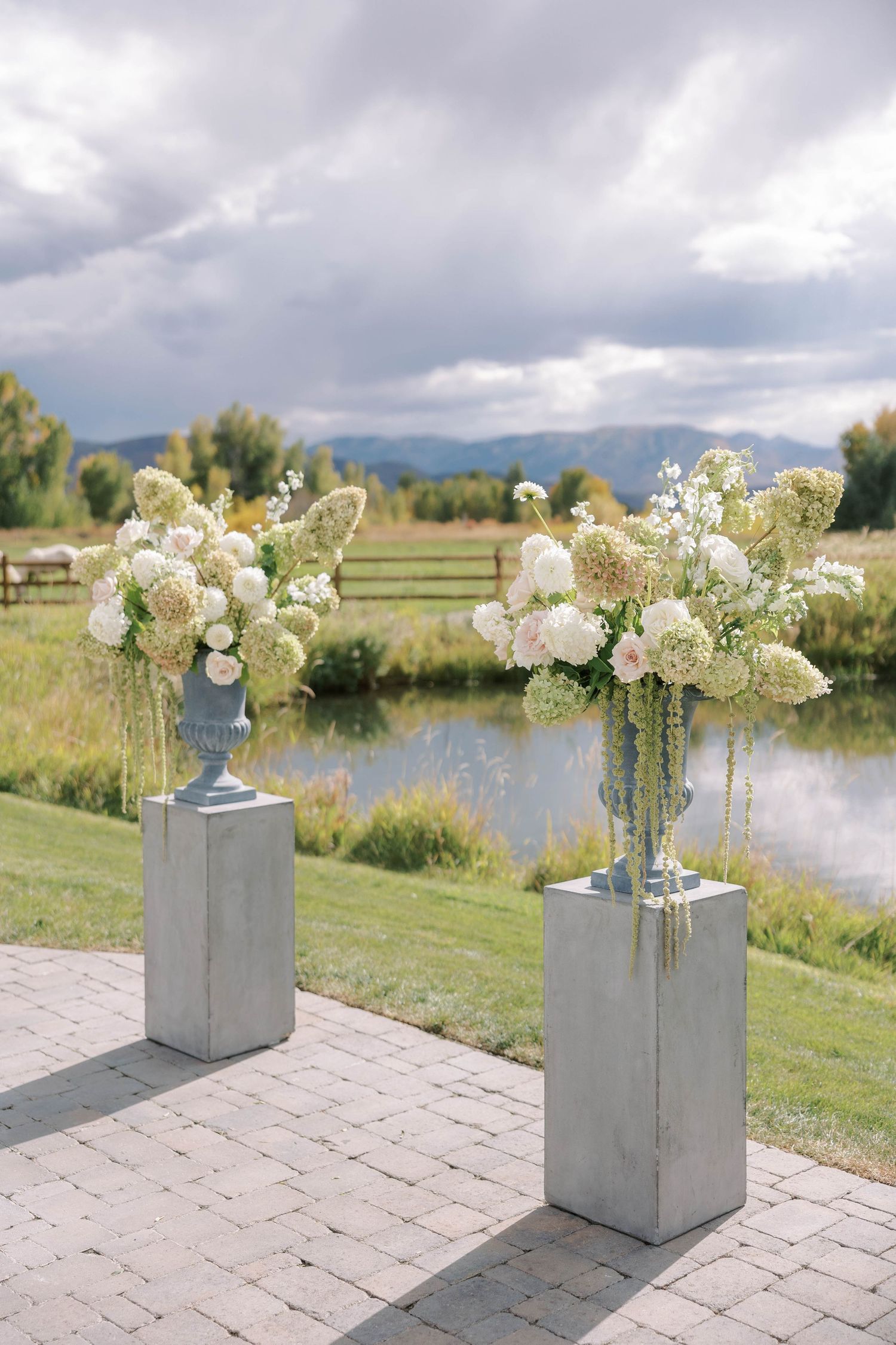 White floral arrangements in concrete planters sit on a stone patio with a scenic mountain lake view in the background.