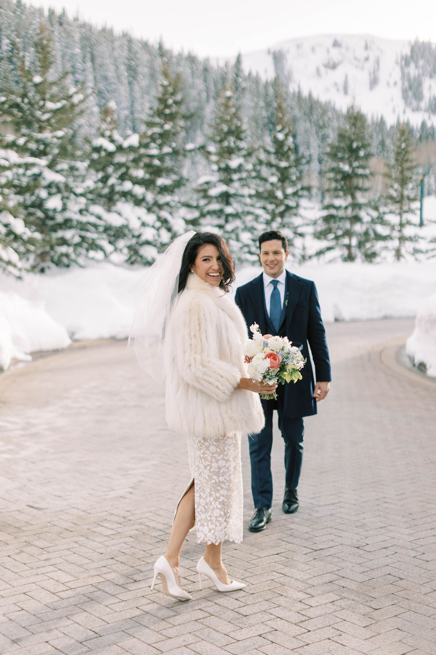 Winter wedding portrait with snow-covered pines and bride in lace dress with fur wrap.