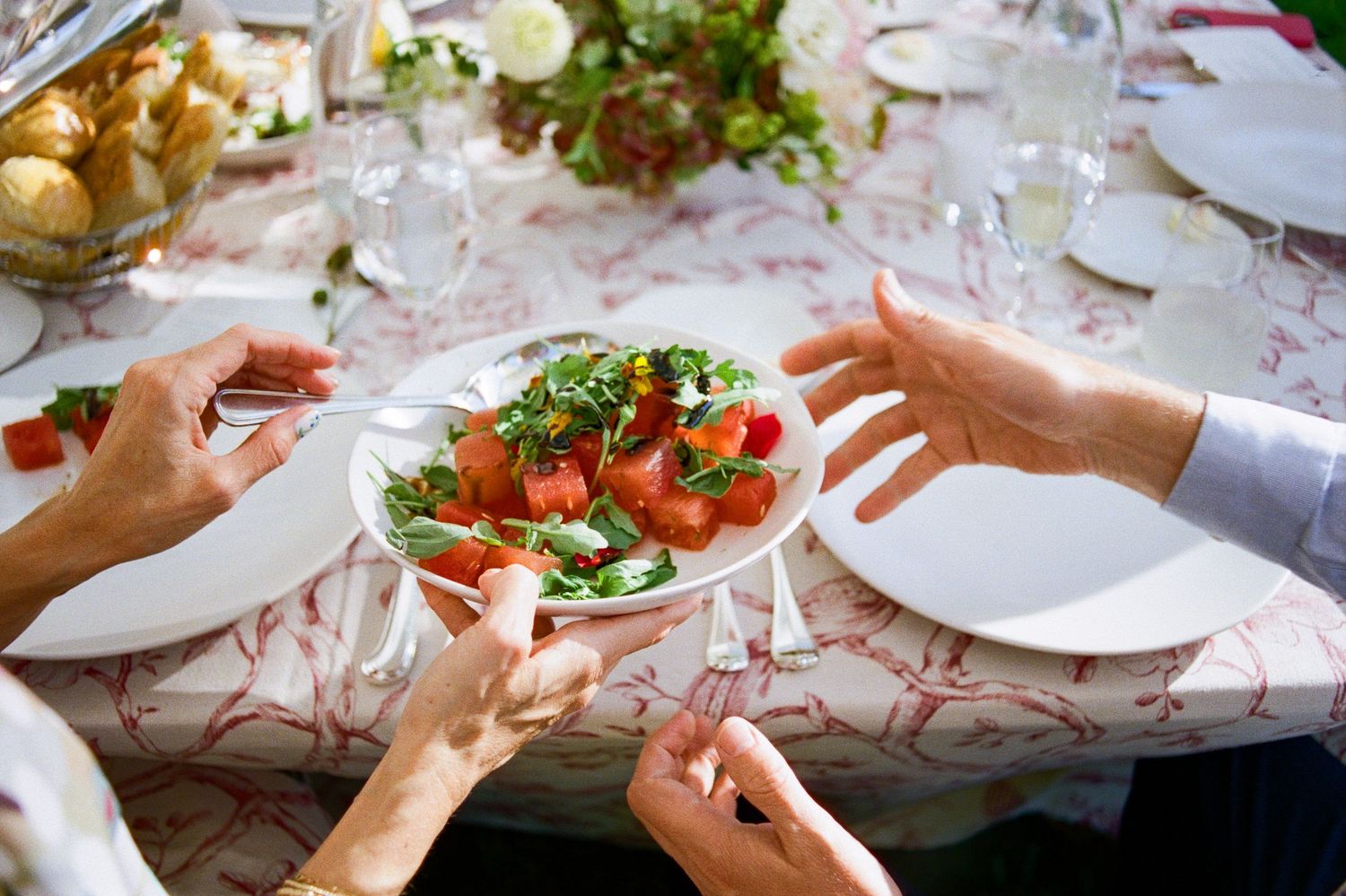 People passing plates and serving food at a dinner party table with white dishes and fresh salads.