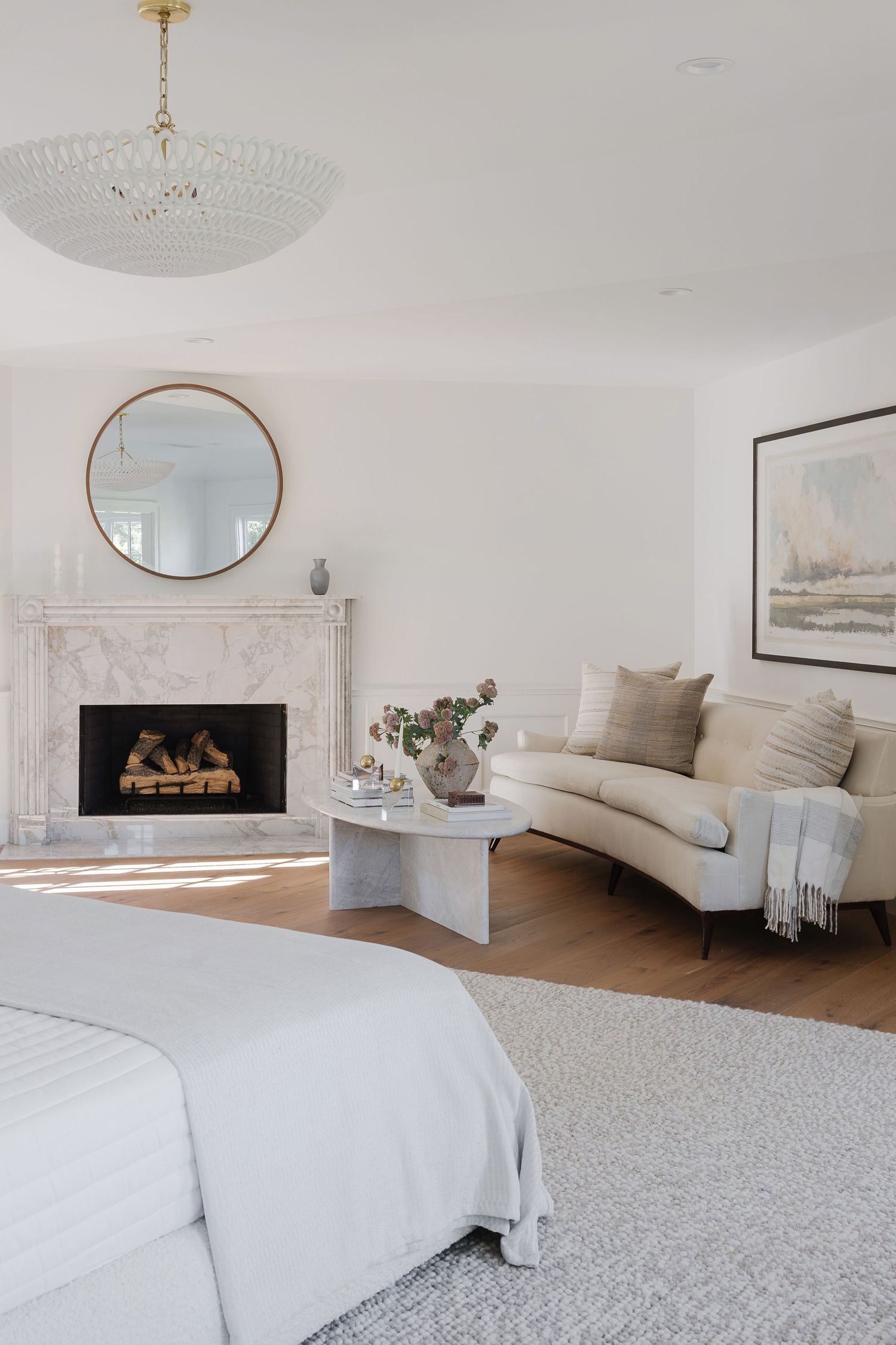 Elegant white bedroom with marble fireplace, round gold mirror, curved beige sofa, crystal chandelier, and neutral decor.