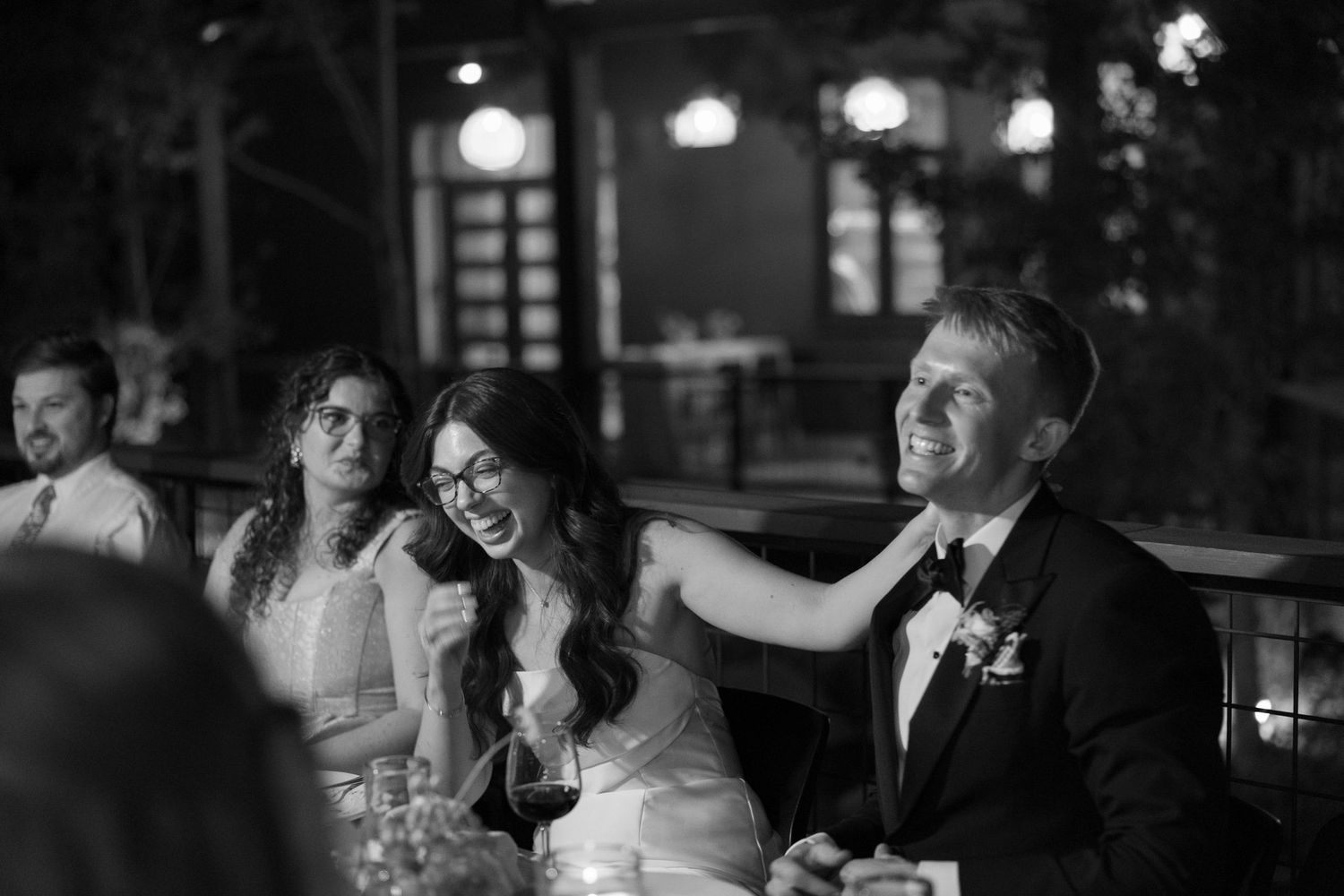 Wedding reception guests share a joyful moment during speeches at an evening celebration in black and white.