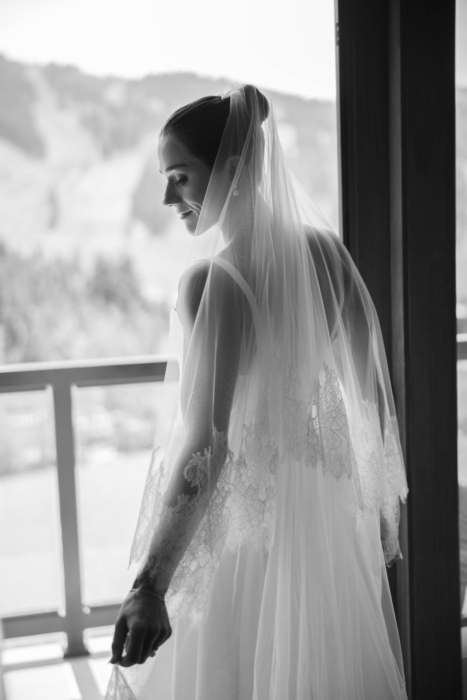 Silhouettes of a bride in her wedding gown and veil against large windows with mountain views.