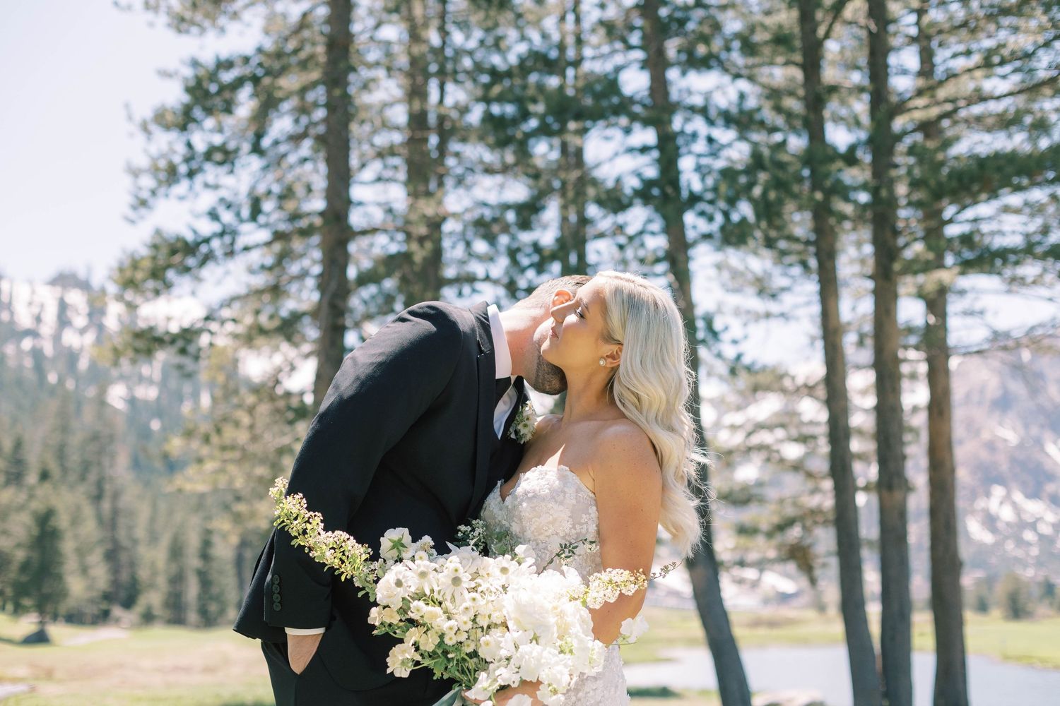 A romantic wedding moment captured in a mountain setting with pine trees and white bouquet.