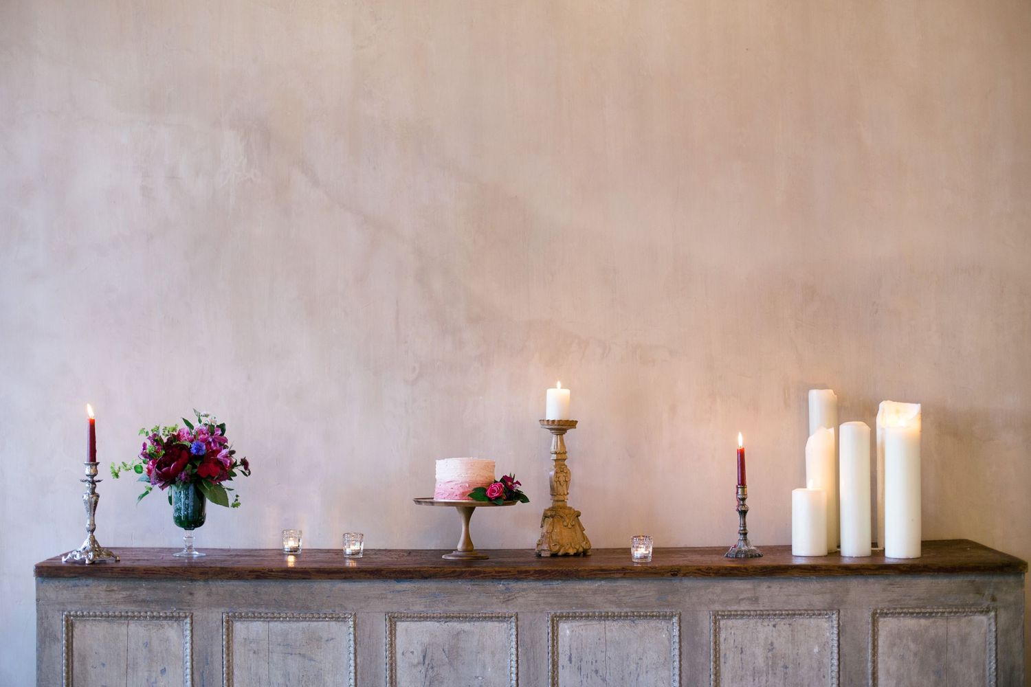 Elegant display of candles and flowers on rustic wooden sideboard against neutral wall.