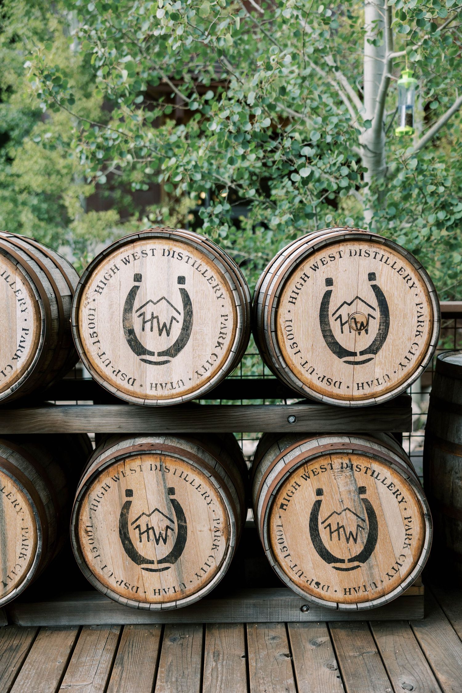 Wooden whiskey barrels with branded logos stacked on wooden deck with trees in background.