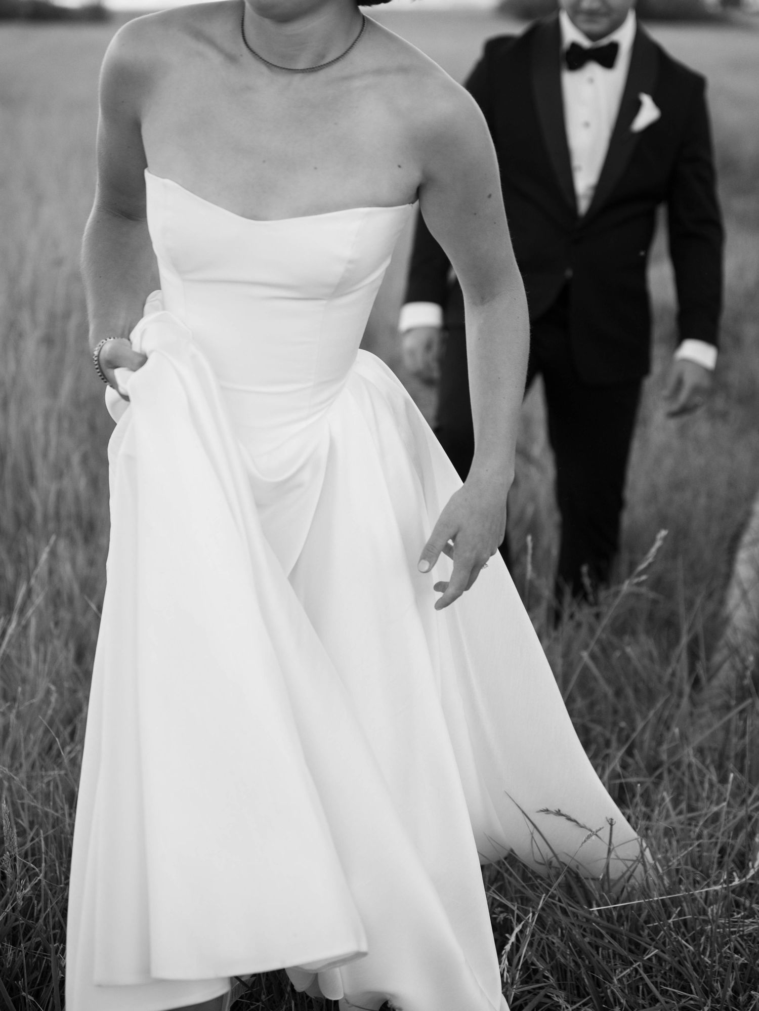 A bride in a white strapless wedding dress walks through a grassy field with her dress flowing in the breeze.