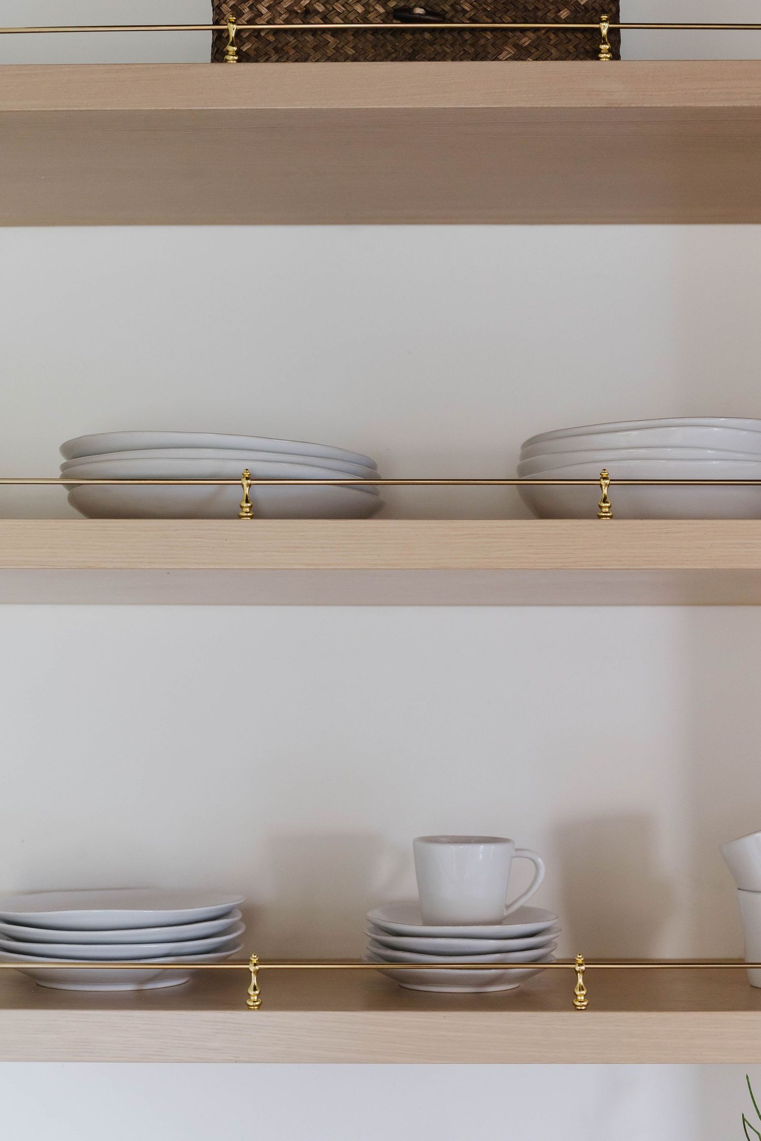 Light wood shelves with gold brackets display stacked white ceramic plates, saucers, and a teacup against a white wall.